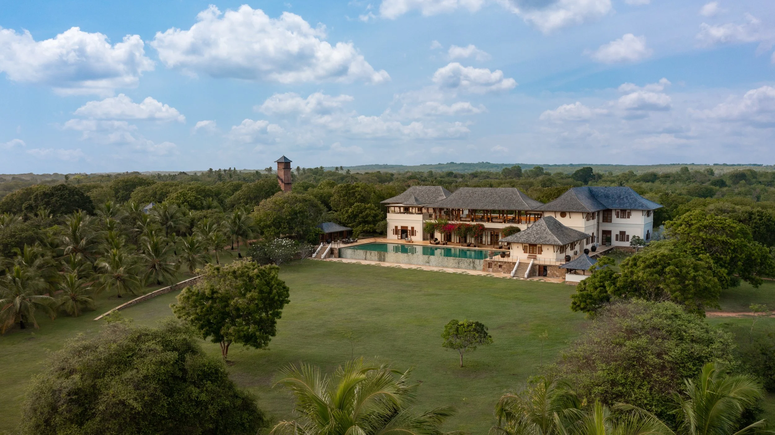 A large luxury house with multiple roofs, a swimming pool, and a lush green lawn surrounded by trees and tropical plants under a partly cloudy sky.
