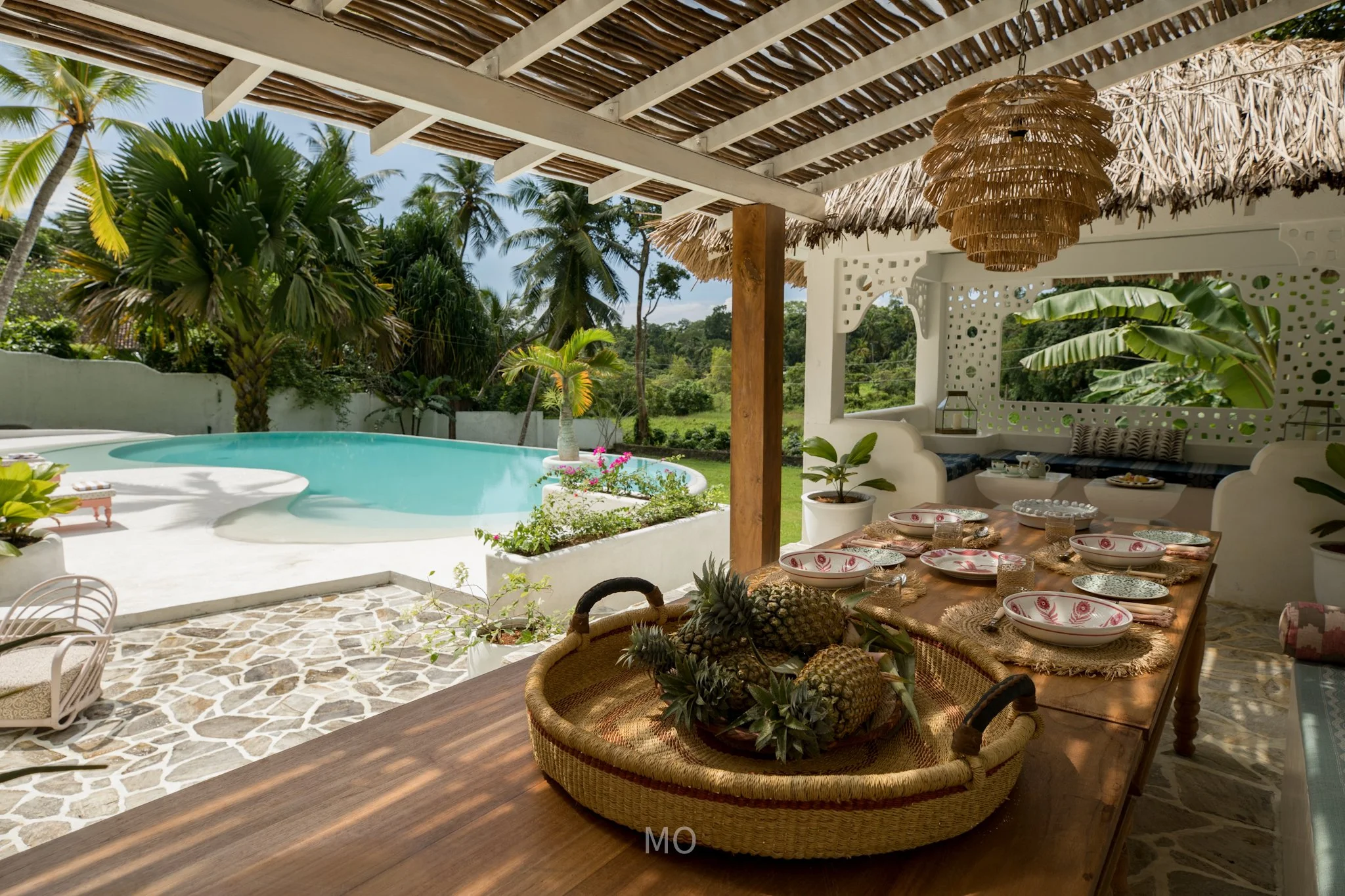 Outdoor patio with a dining table set for a meal, decorated with pineapples in a basket, overlooking a swimming pool surrounded by tropical palm trees.