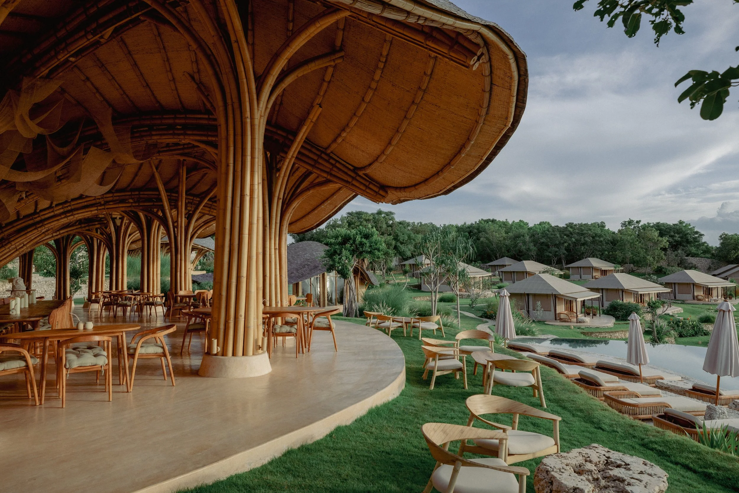 A tropical resort with a bamboo-style open-air restaurant on a curved deck, overlooking a lush landscape with small bungalow-style accommodations and a swimming pool, under a cloudy sky.