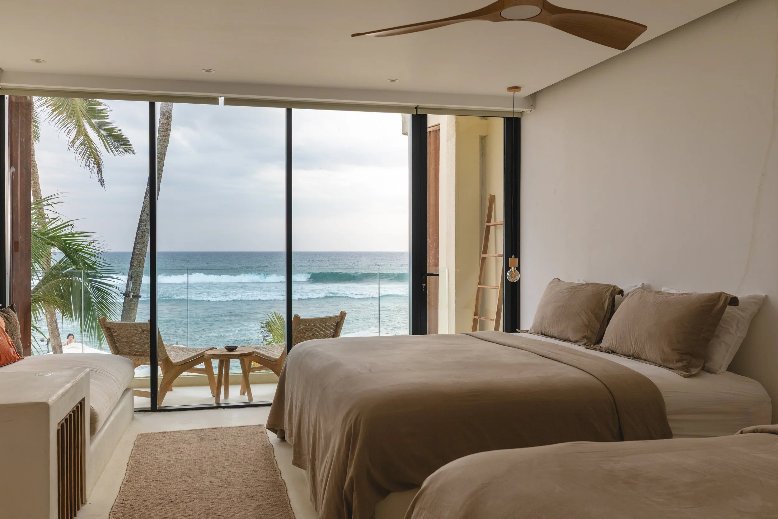 A bedroom with two beds, beige linens, and pillows, overlooking a balcony with a view of the ocean and waves, through glass sliding doors.