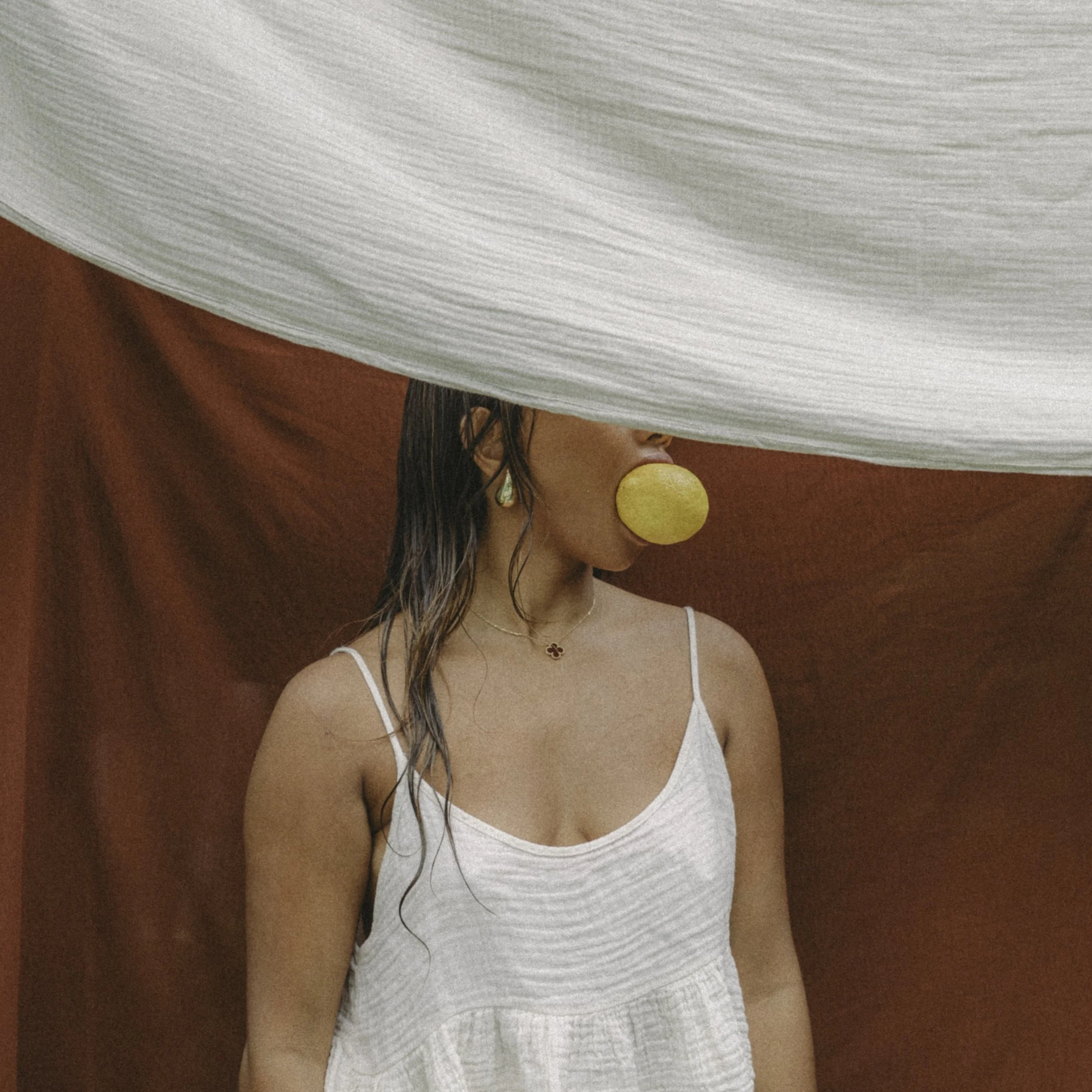 A woman with wet hair blowing a yellow bubble gum, wearing a white sleeveless top, standing against a red background with a large white fabric overhead.