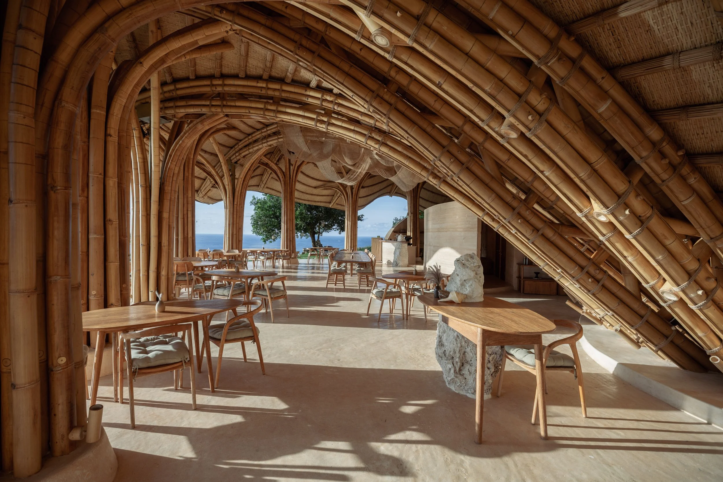 Interior of a thatched roof restaurant with wooden tables and chairs, large open archways revealing a view of the ocean and a tree outside.