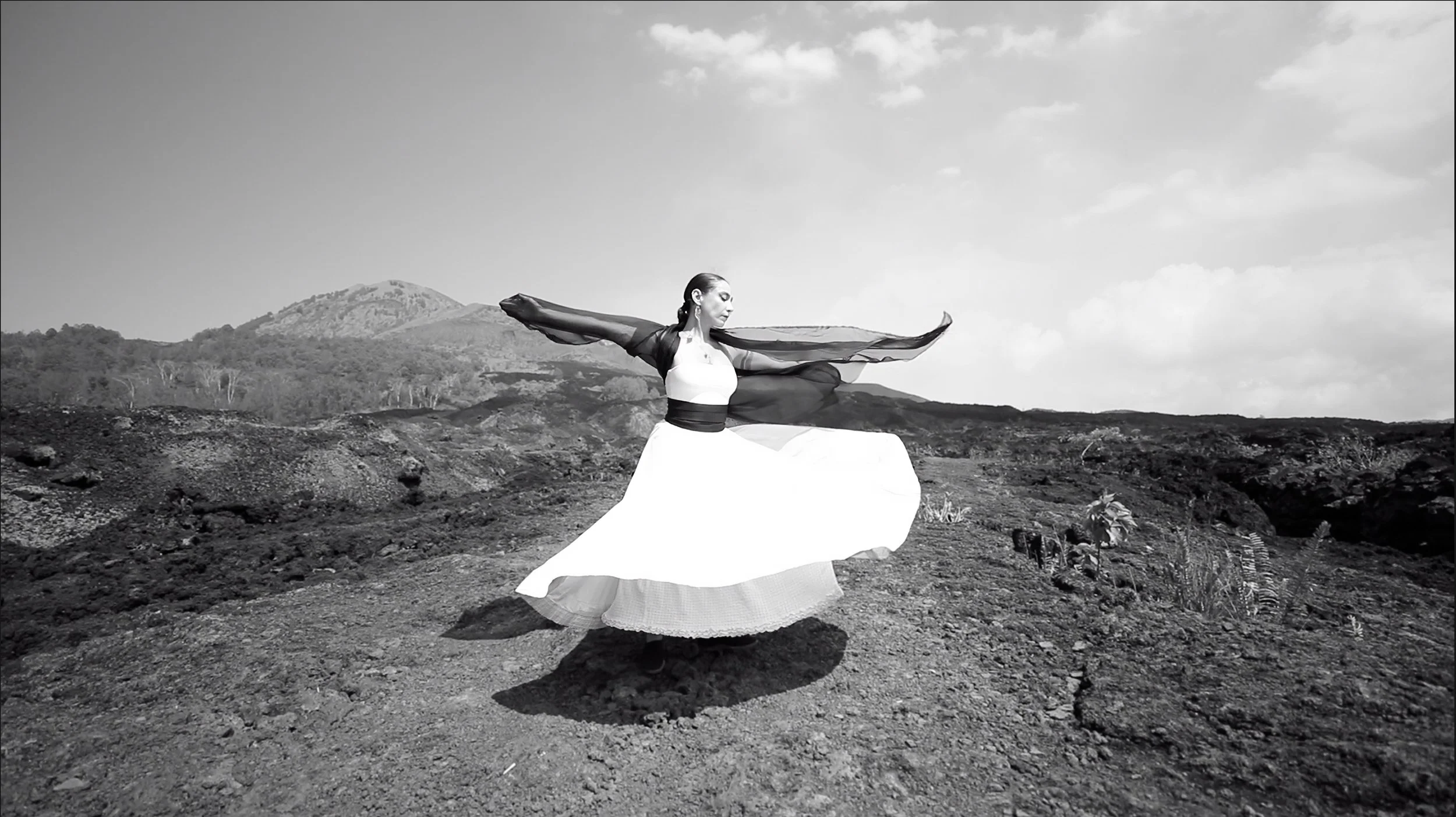A woman in a flowing dress is spinning in a barren outdoor landscape with mountains in the background, captured in black and white.
