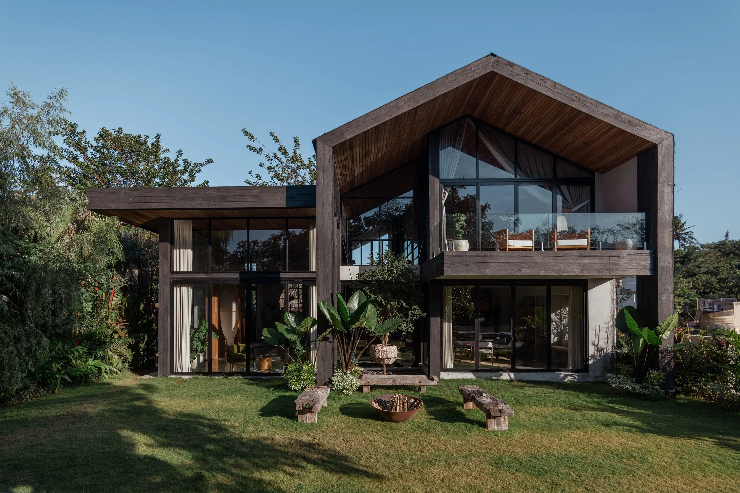 Modern two-story house with large glass windows and wooden accents, surrounded by a green lawn and trees.