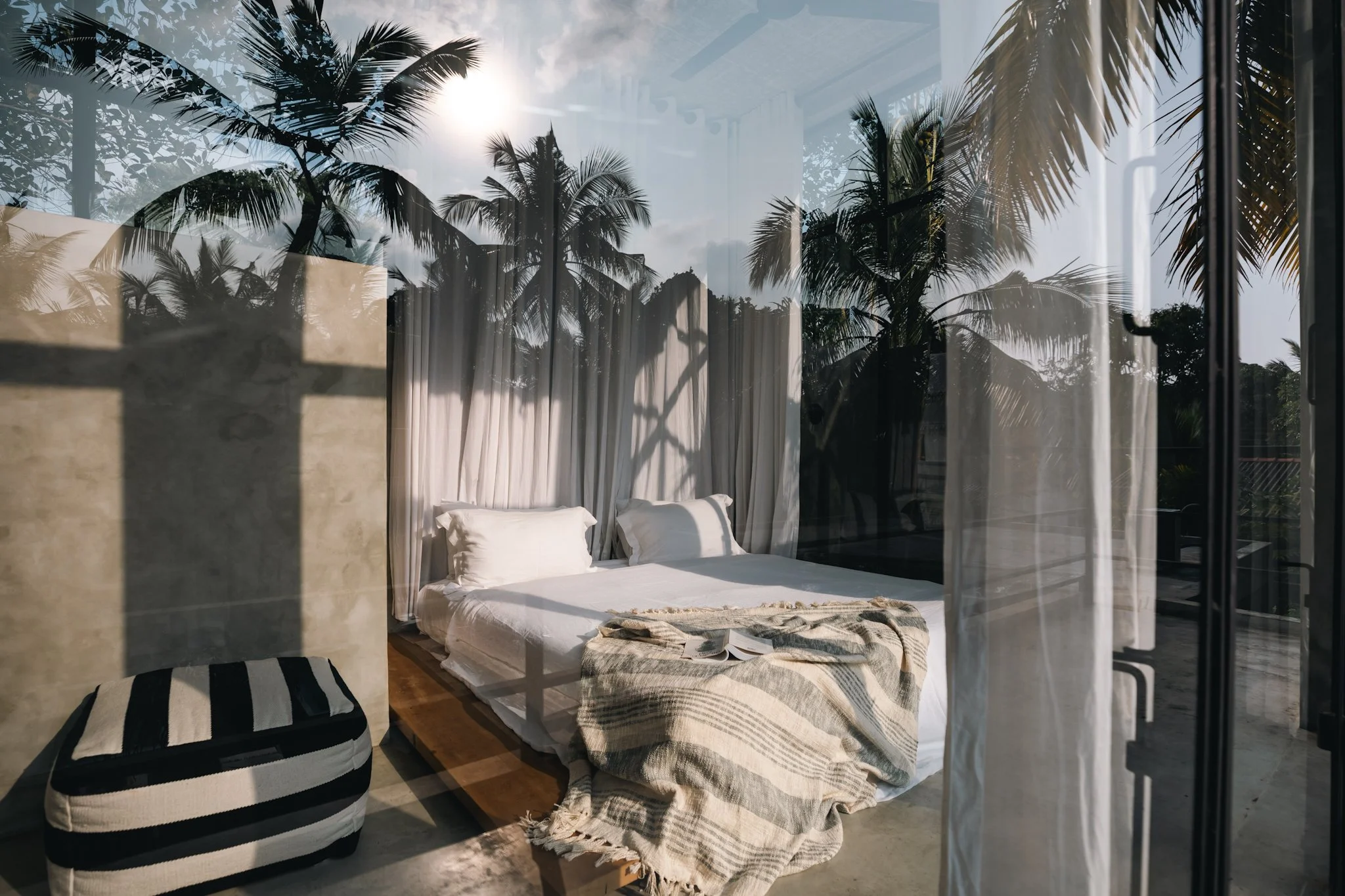 A cozy bedroom with a king-sized bed, white linens, and pillows, reflected in a glass window showing palm trees and sunlight outside, with a striped ottoman at the foot of the bed and sheer curtains.