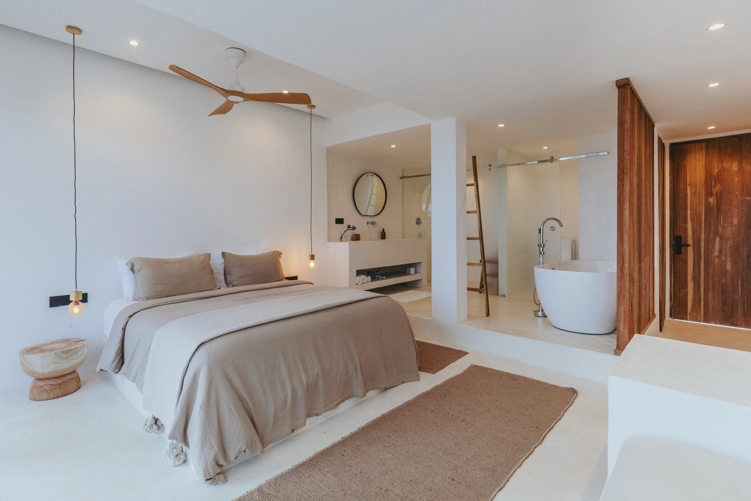 A minimalist bedroom with a beige bed, wooden side tables, two hanging pendant lights, and a ceiling fan. An open bathroom area features a white bathtub, a ladder, and a wooden sliding door.