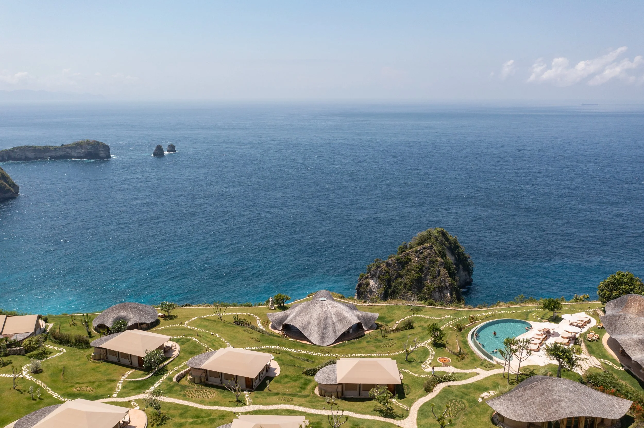 Aerial view of a coastal resort with grass areas, thatched roof cottages, a swimming pool, and ocean waves with rocky islands in the background.