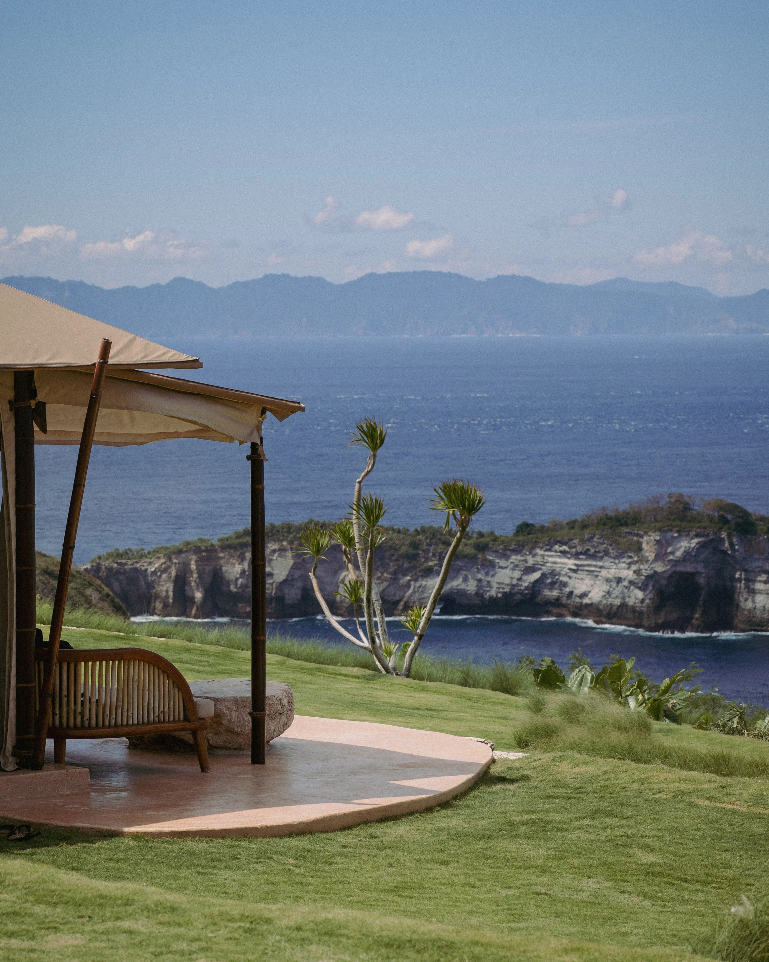A scenic seaside view with a grassy lawn, a beige umbrella, a wooden bench, a small tree, rocky cliffs, and an ocean under a blue sky with clouds.