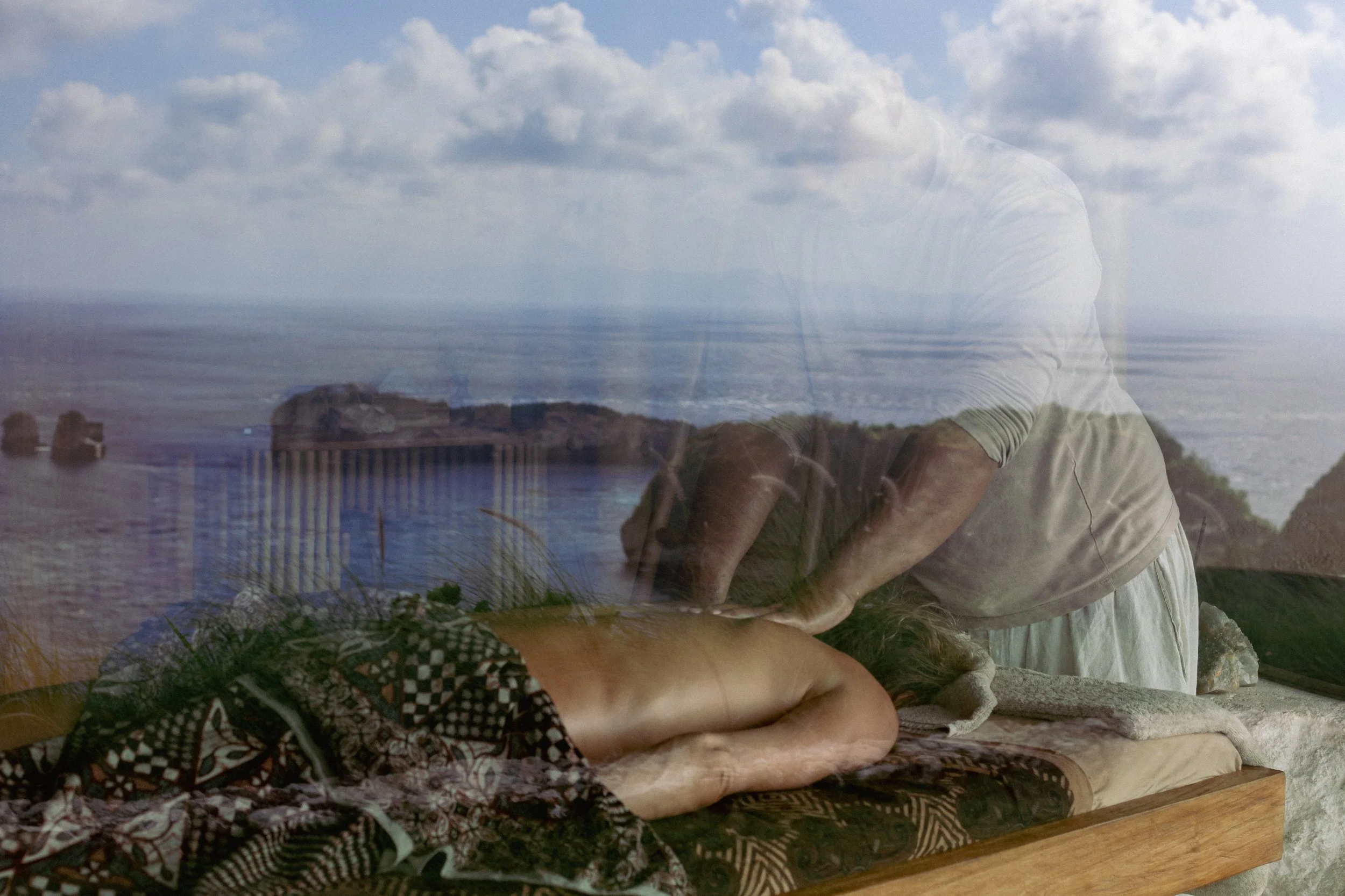 A person receiving a massage outdoors with the ocean, rocks, and a cloudy sky in the background.