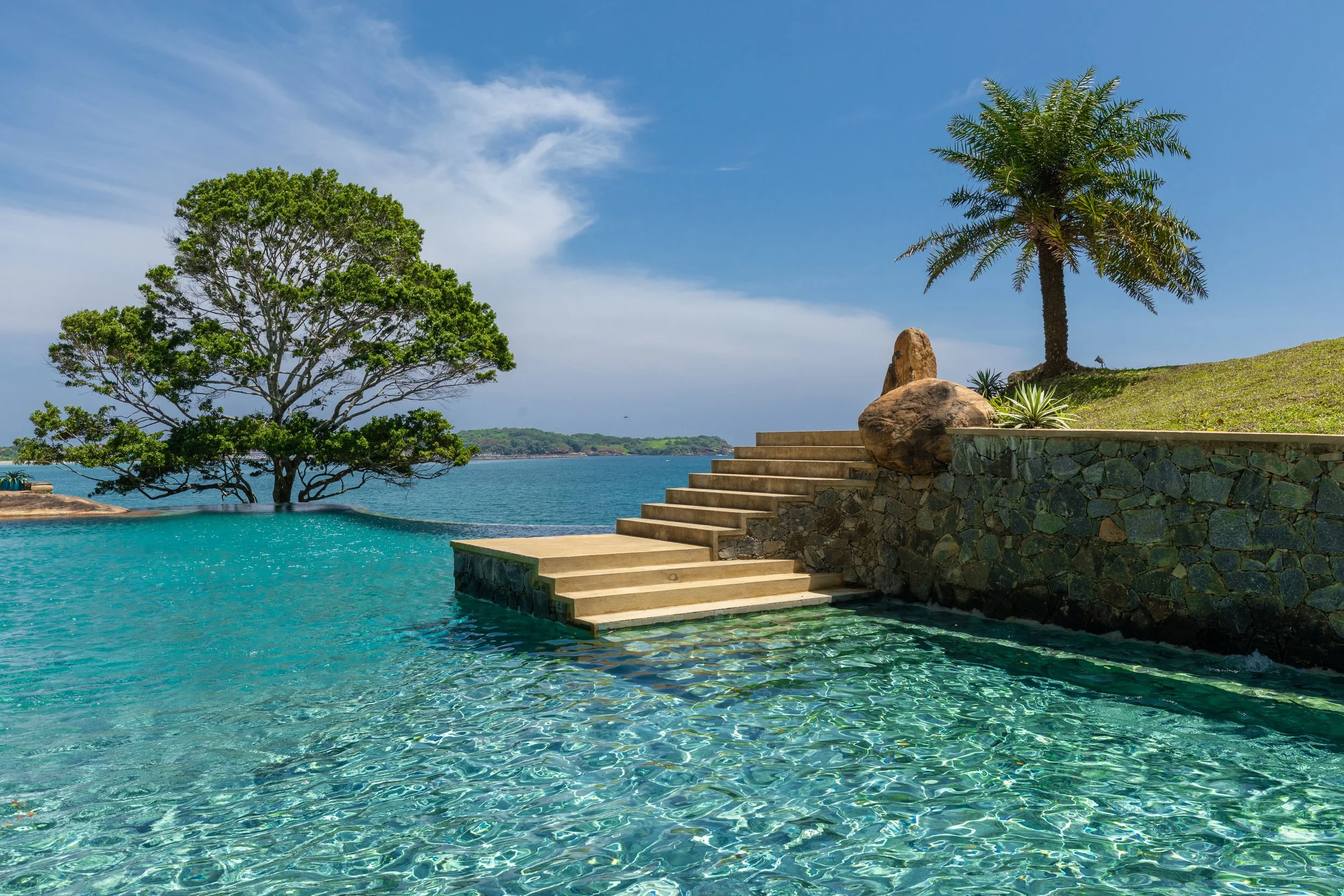 A luxurious infinity pool overlooking the ocean, with steps leading into the water, surrounded by green trees, palm trees, rocks, and a stone wall under a blue sky.