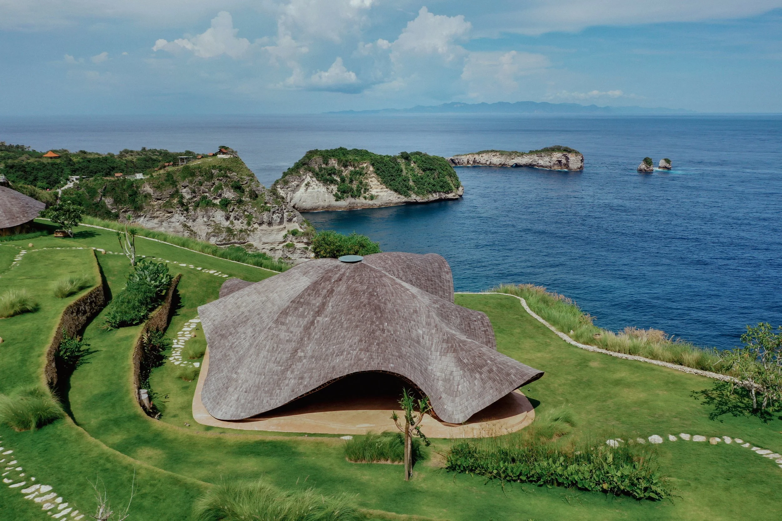Aerial view of a modern wooden building with curved roof, lush green lawn, and pathway, overlooking the ocean with cliffs and small islands in the distance.