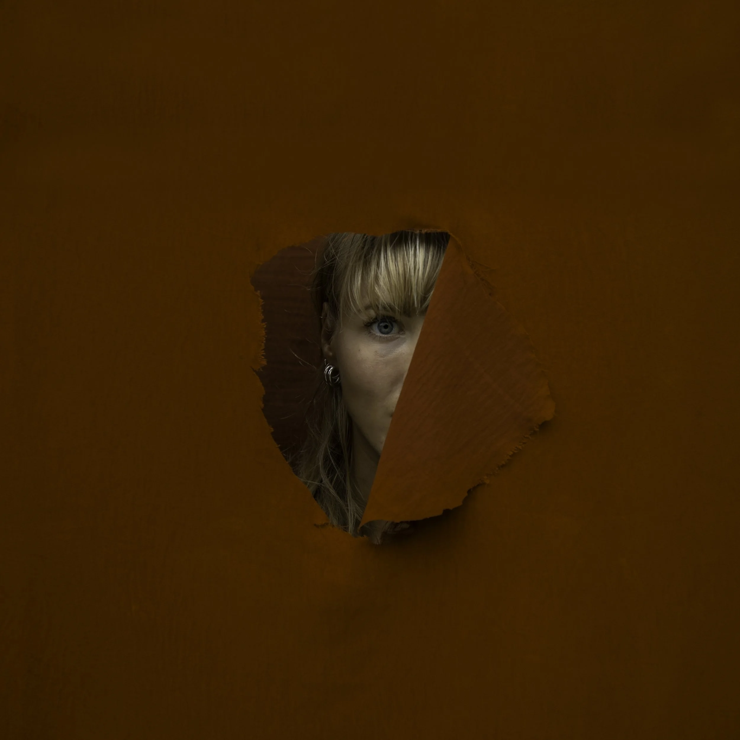 A woman with blonde hair and blue eyes looking through a torn orange paper or cardboard.