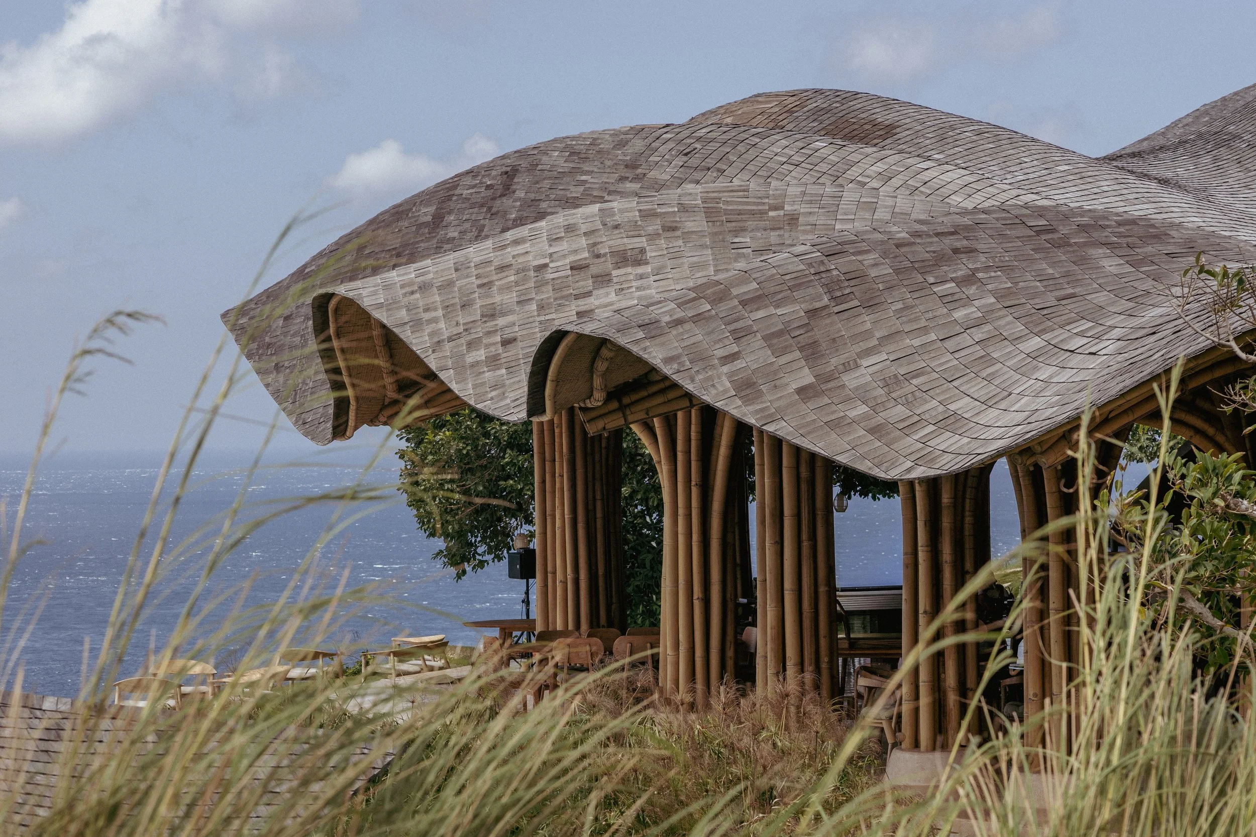 A seaside restaurant or cafe with a bamboo structure and a curved, wooden shingle roof, overlooking the ocean and partly obscured by tall grass in the foreground.