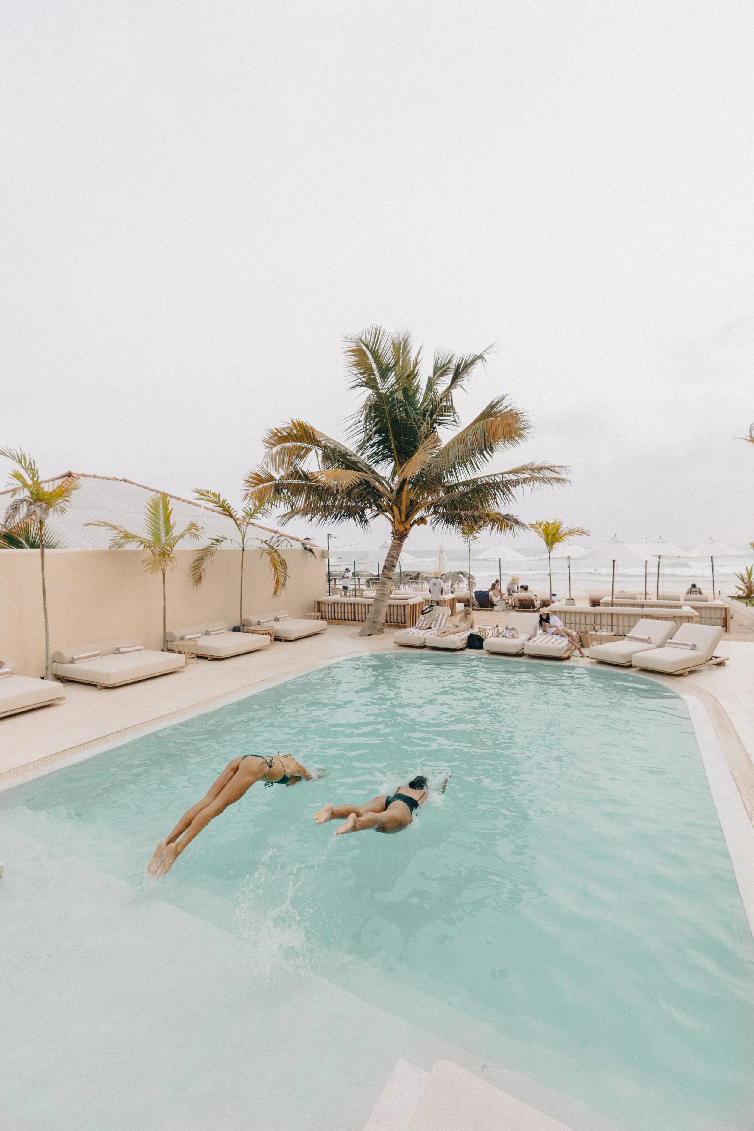 People swimming in a pool with palm trees and beach chairs in the background at a beach resort.