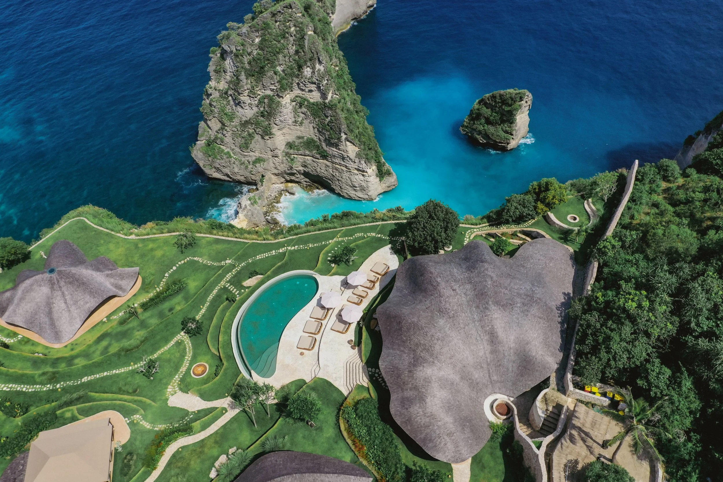 Aerial view of a tropical resort with thatched roofs, a swimming pool, lush green landscaping, and a coastline with steep cliffs and blue ocean water.