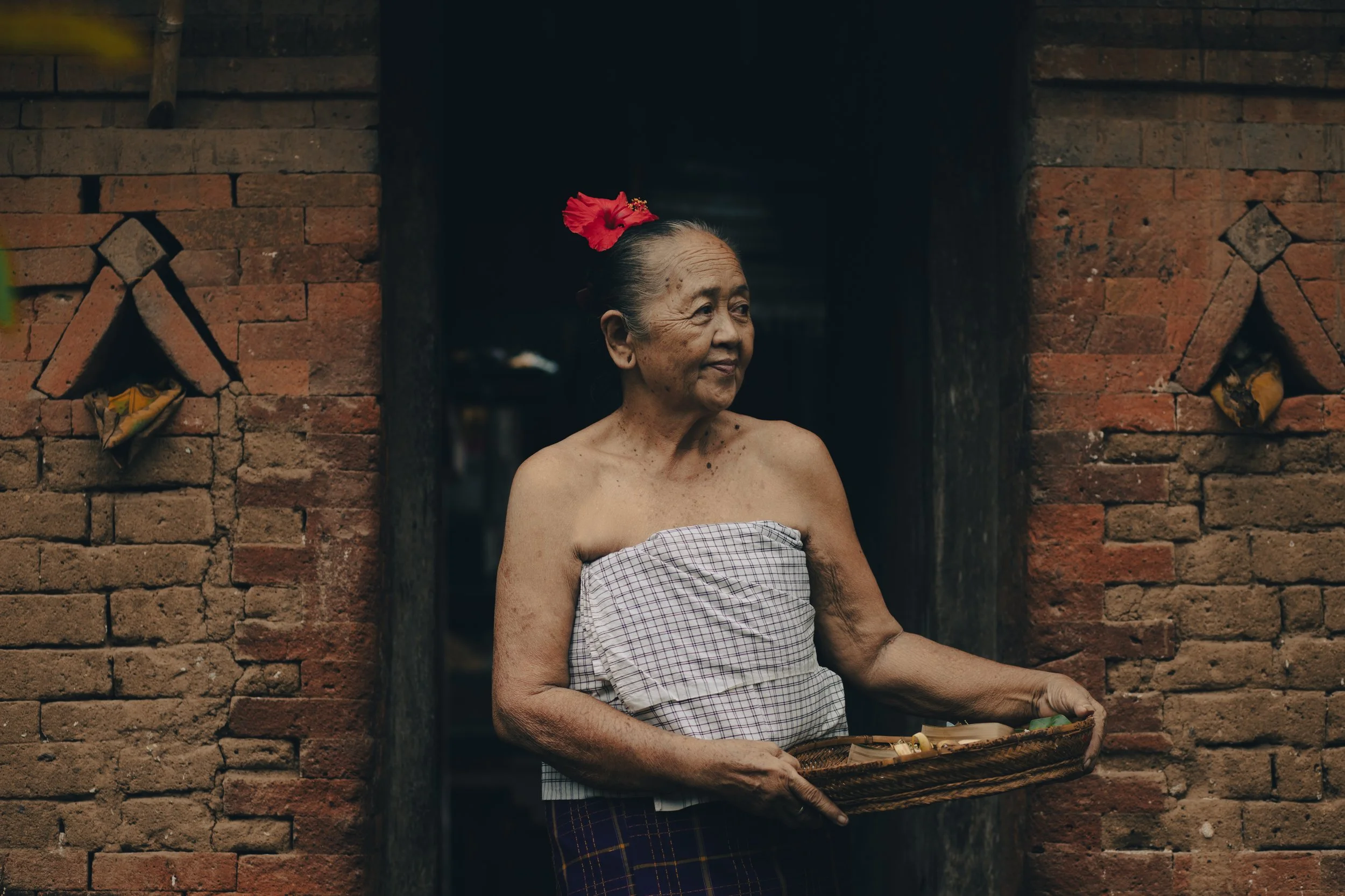 An elderly woman with tan skin and gray hair, wearing a piece of cloth wrapped around her chest and a purple checkered skirt, holding a basket and standing in a doorway of a brick building, with a red flower in her hair.