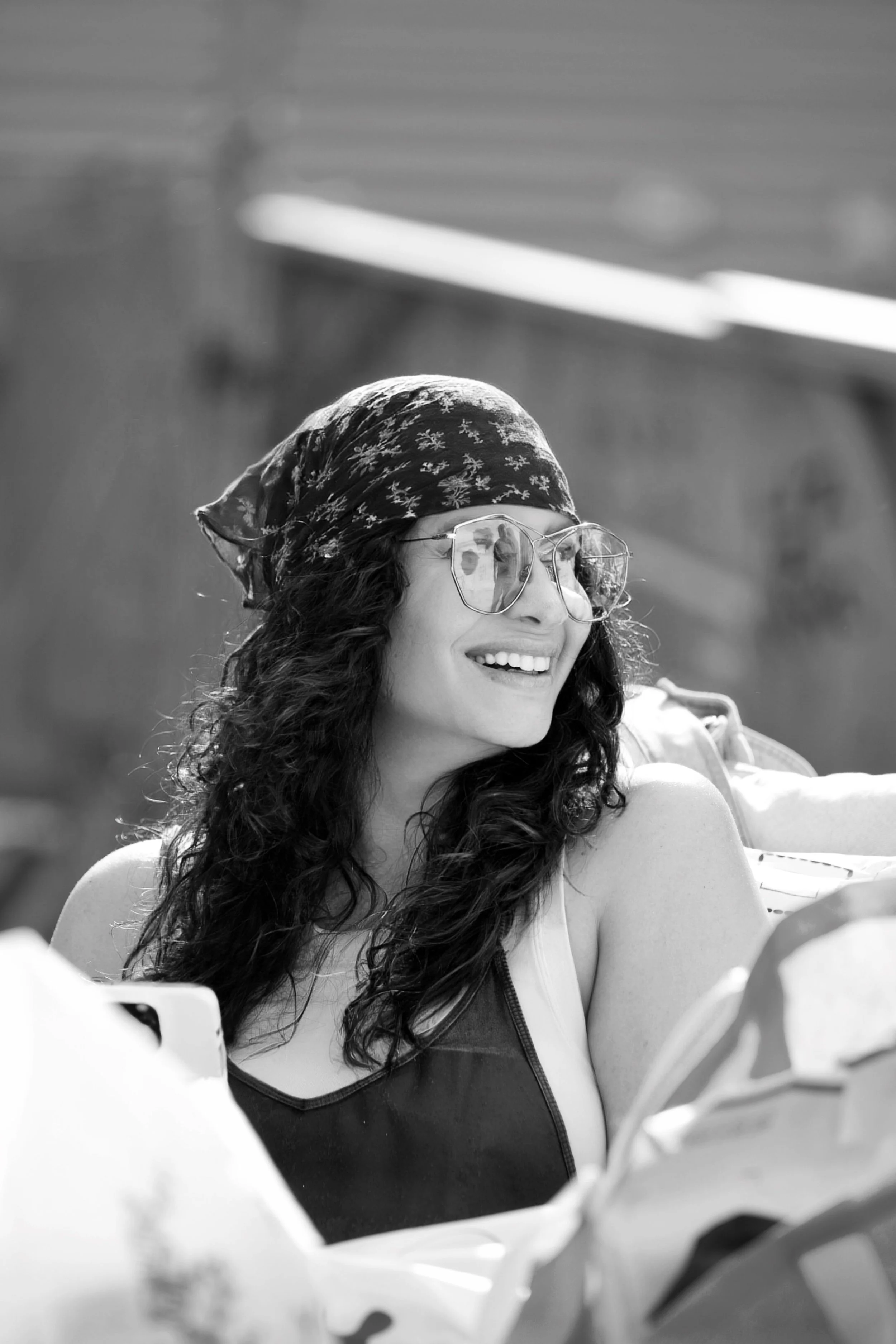 A woman with curly hair wearing glasses, a headscarf, and a sleeveless top, smiling outdoors in sunlight.