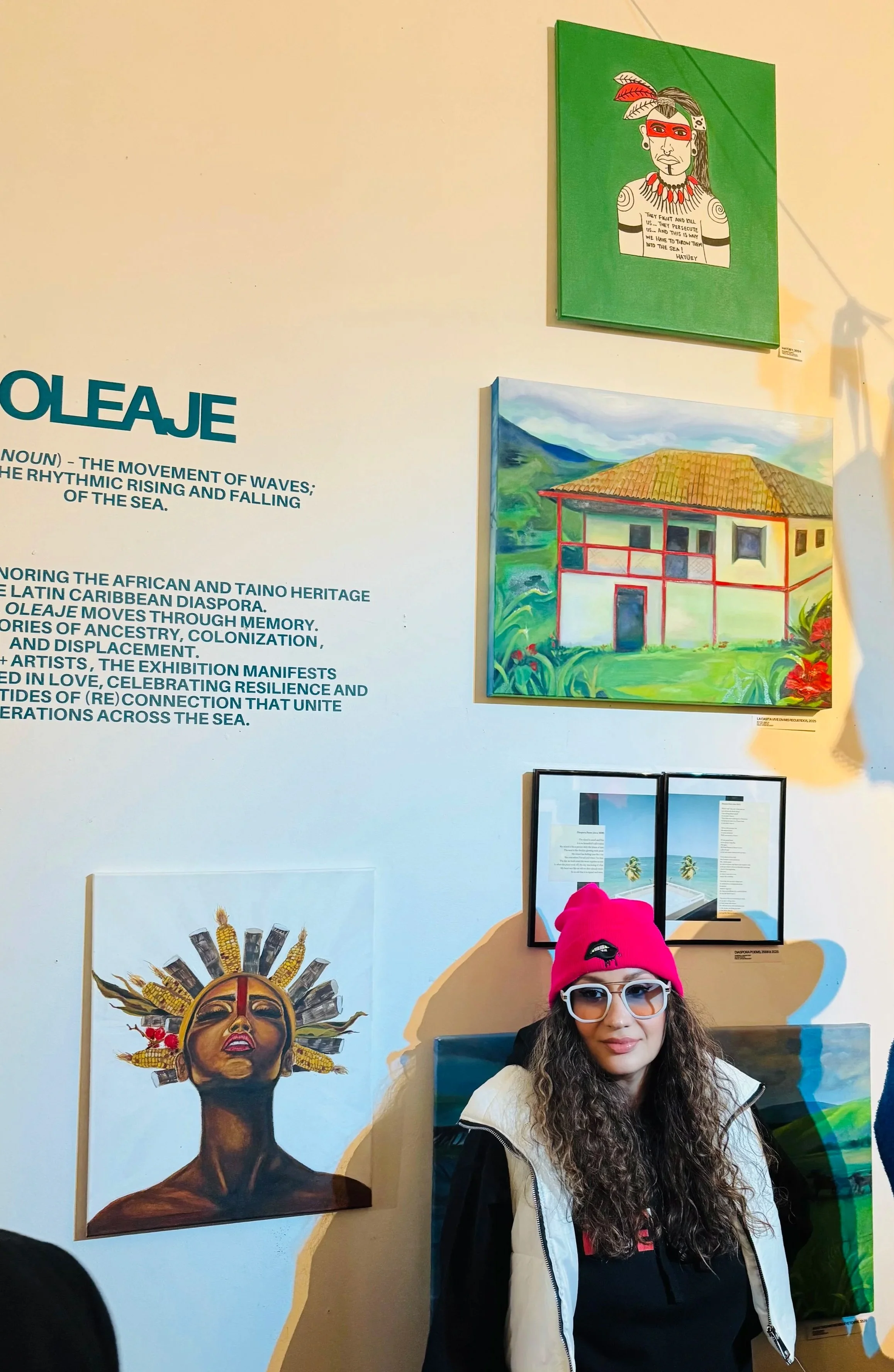A young woman with long curly hair, glasses, and a bright pink beanie hat, standing in front of an art display wall. The wall features several paintings, including a portrait of a person with feathers and jewelry, a colorful house, and a woman with an elaborate headdress. There is also a section of blue text explaining the art exhibit.