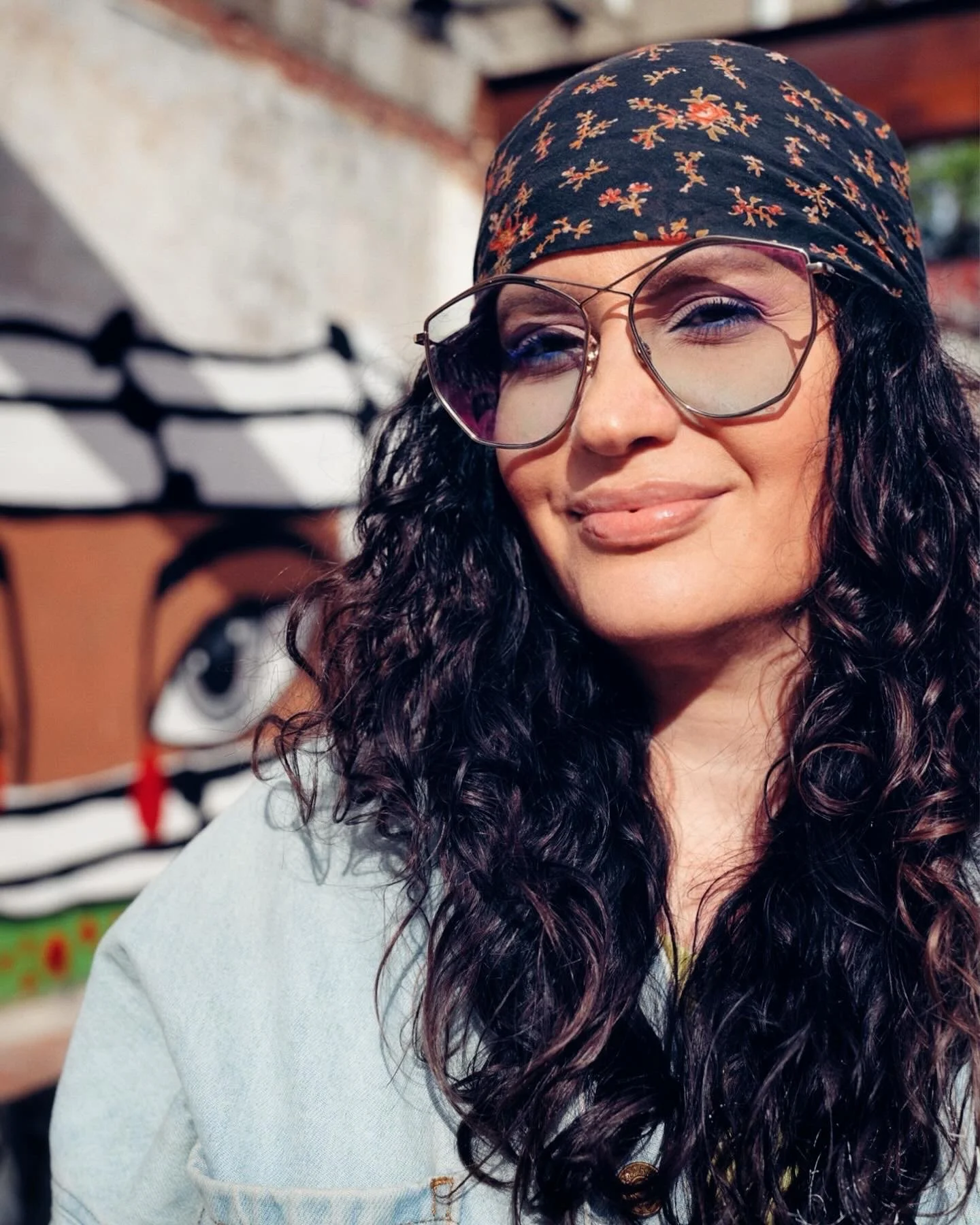 Smiling woman with long curly dark hair, wearing large glasses, a floral bandana, and a light-colored denim jacket, outdoors with graffiti art in the background.