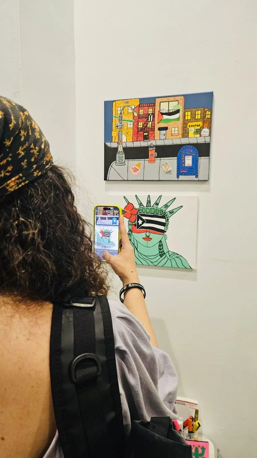 A woman taking a photo of two artwork pieces on a gallery wall. One piece features a colorful city street scene, the other a stylized Statue of Liberty with the Puerto Rican flag across her eyes.