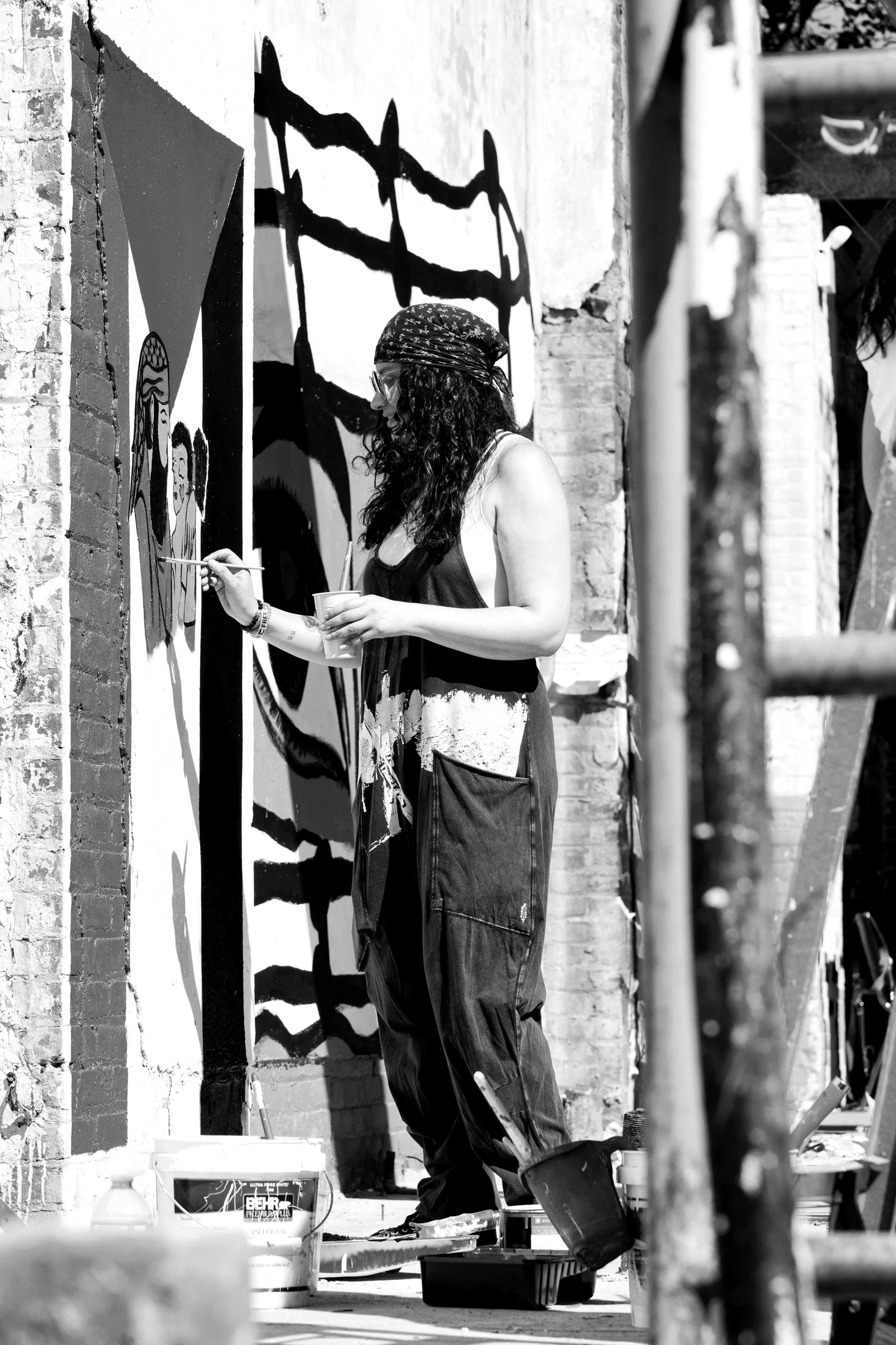 A woman with curly hair, wearing a bandana and glasses, is painting a mural on a brick wall outdoors. She is holding a cup in one hand and a paintbrush in the other, and standing on a platform with painting supplies around her.