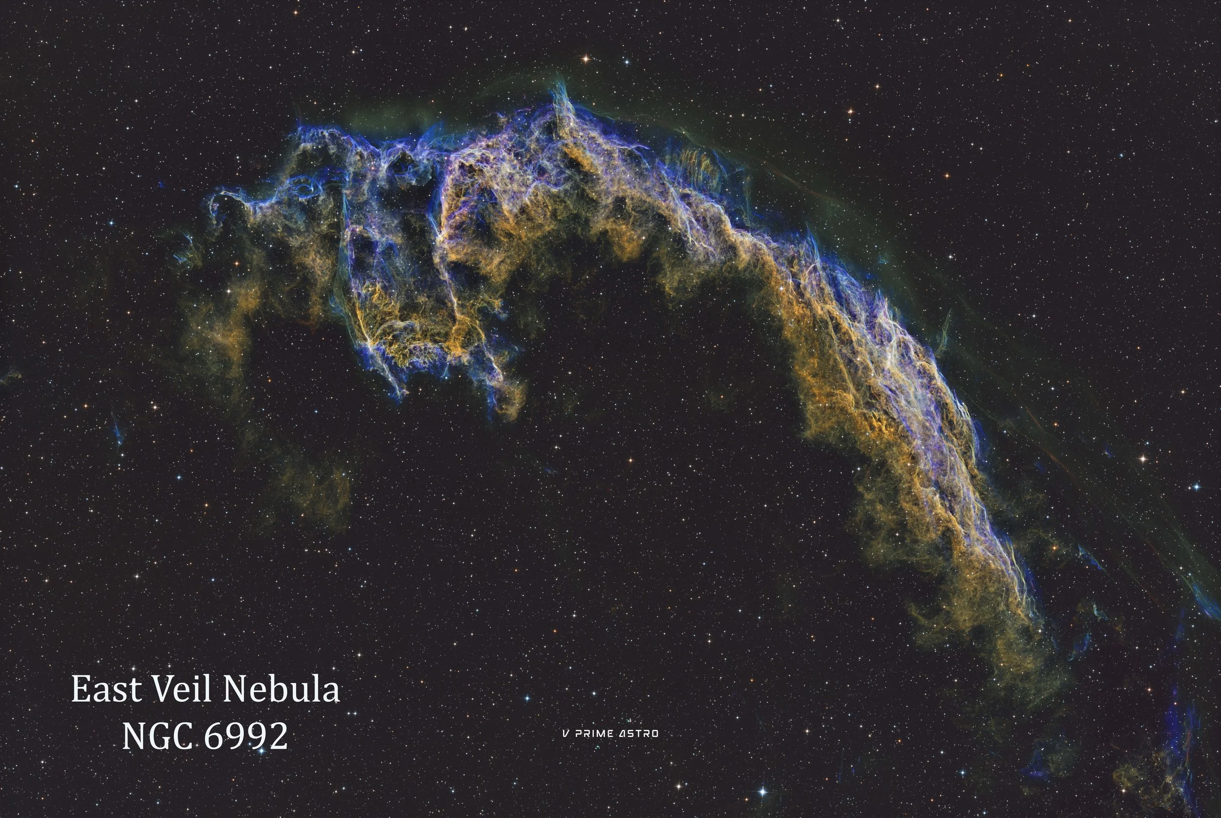 Colorful image of the East Veil Nebula, a supernova remnant, with stars scattered across the background. The nebula features intricate filaments of gas in blue, yellow, and green hues. Text at the bottom left reads 'East Veil Nebula NGC 6992', and at