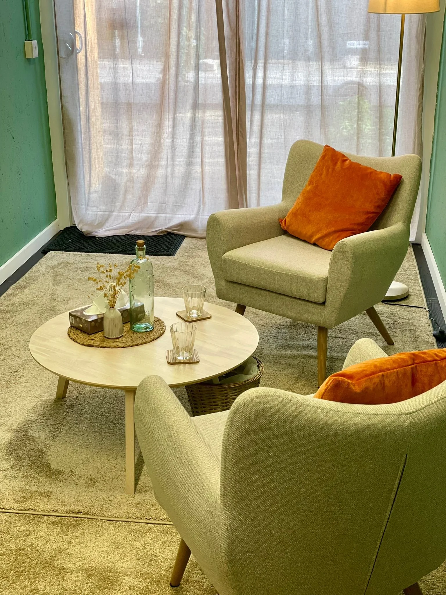 Private and light therapy room with two armchairs, a round wooden coffee table with decorative items, a beige carpet, a floor lamp, and a beige curtain covering a window with sunlight coming through.