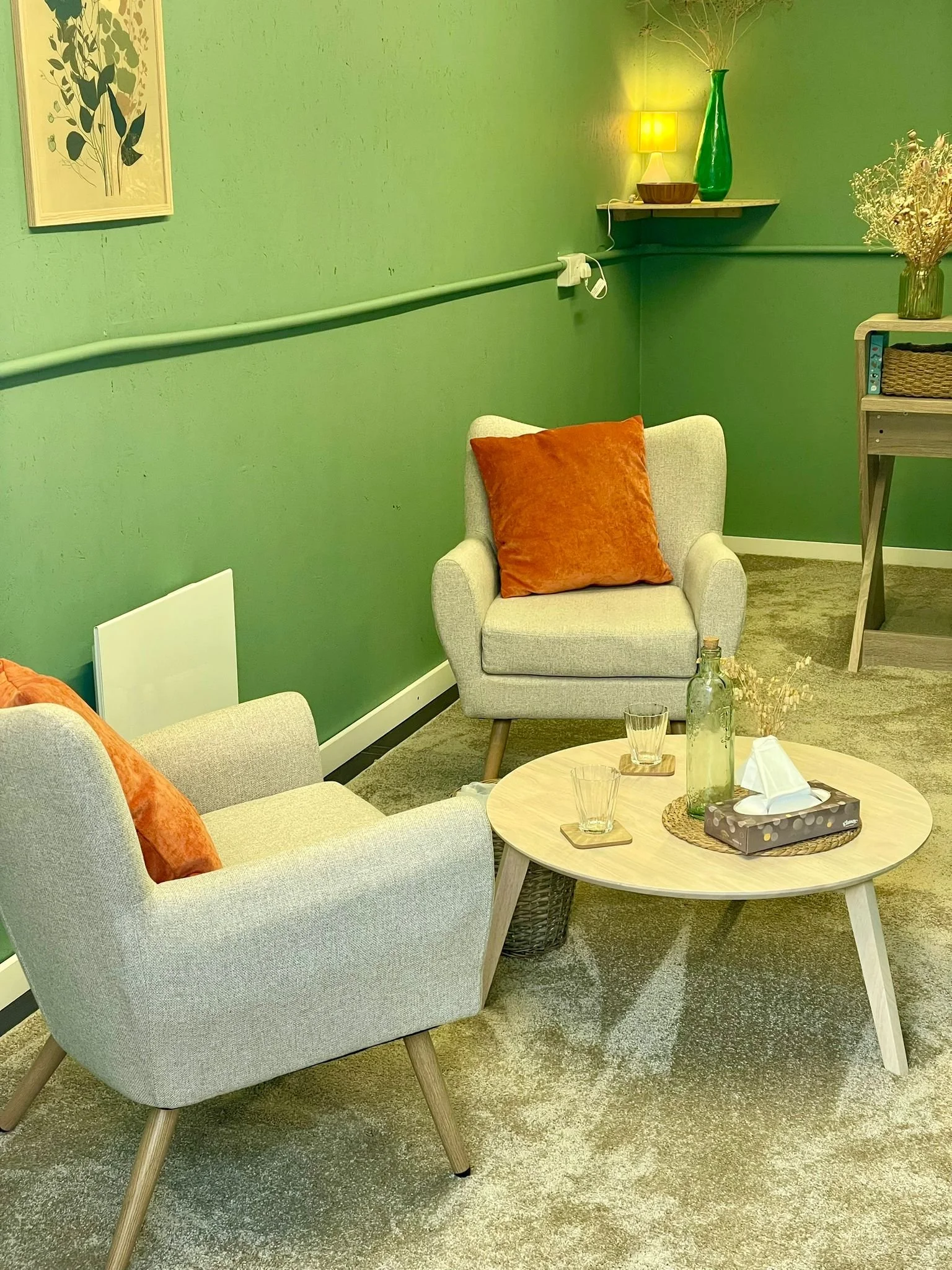 Therapy room with two grey armchairs, both with orange cushions, around a round wooden table with glasses, a bottle, tissues, and decorative items. A green wall with a picture, shelf with a lamp and vases, and a desk with a basket and dried flowers.