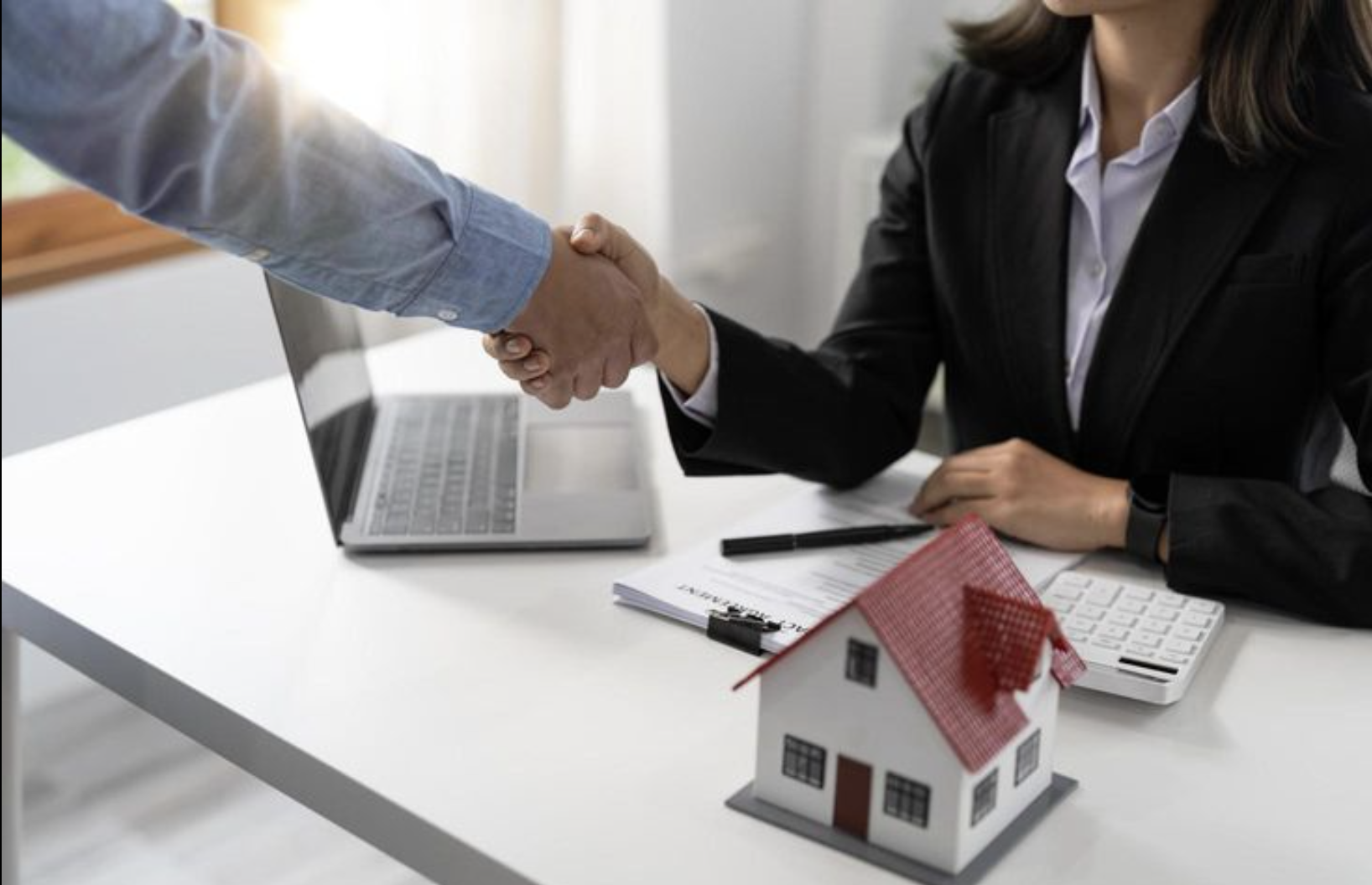 Two people shaking hands across an office desk, with a model house, a laptop, and documents on the desk.