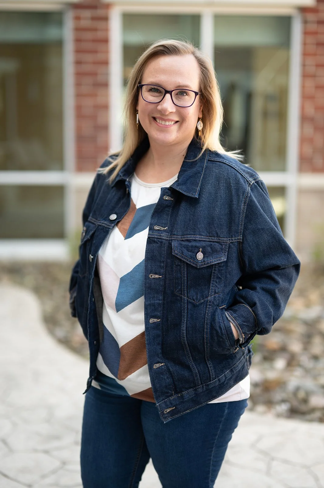 A woman with blonde hair, glasses, and a purple patterned blouse standing outdoors near a tree, smiling at the camera.