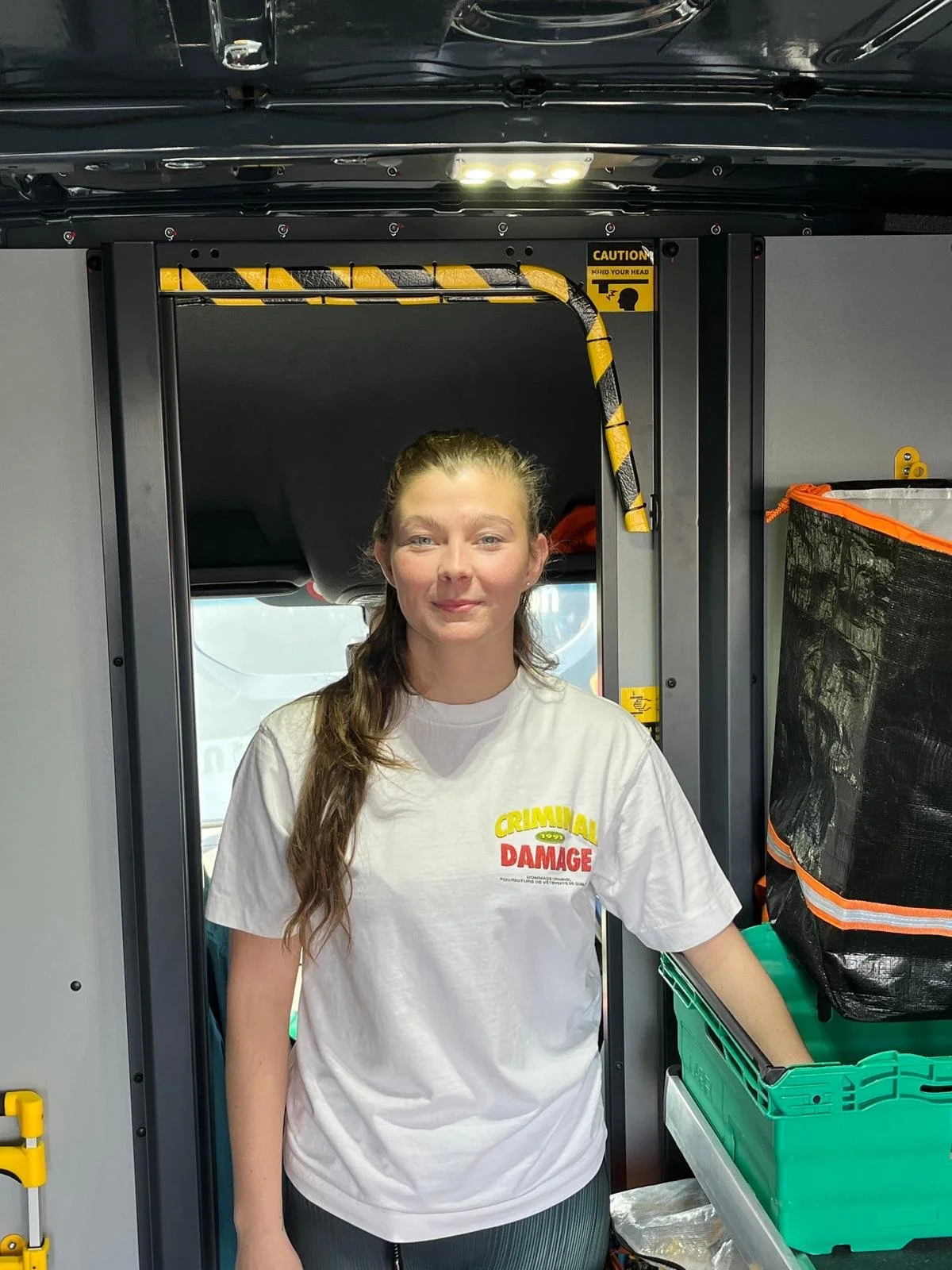 A young woman with long brown hair, wearing a white T-shirt, stands inside a courier vehicle.