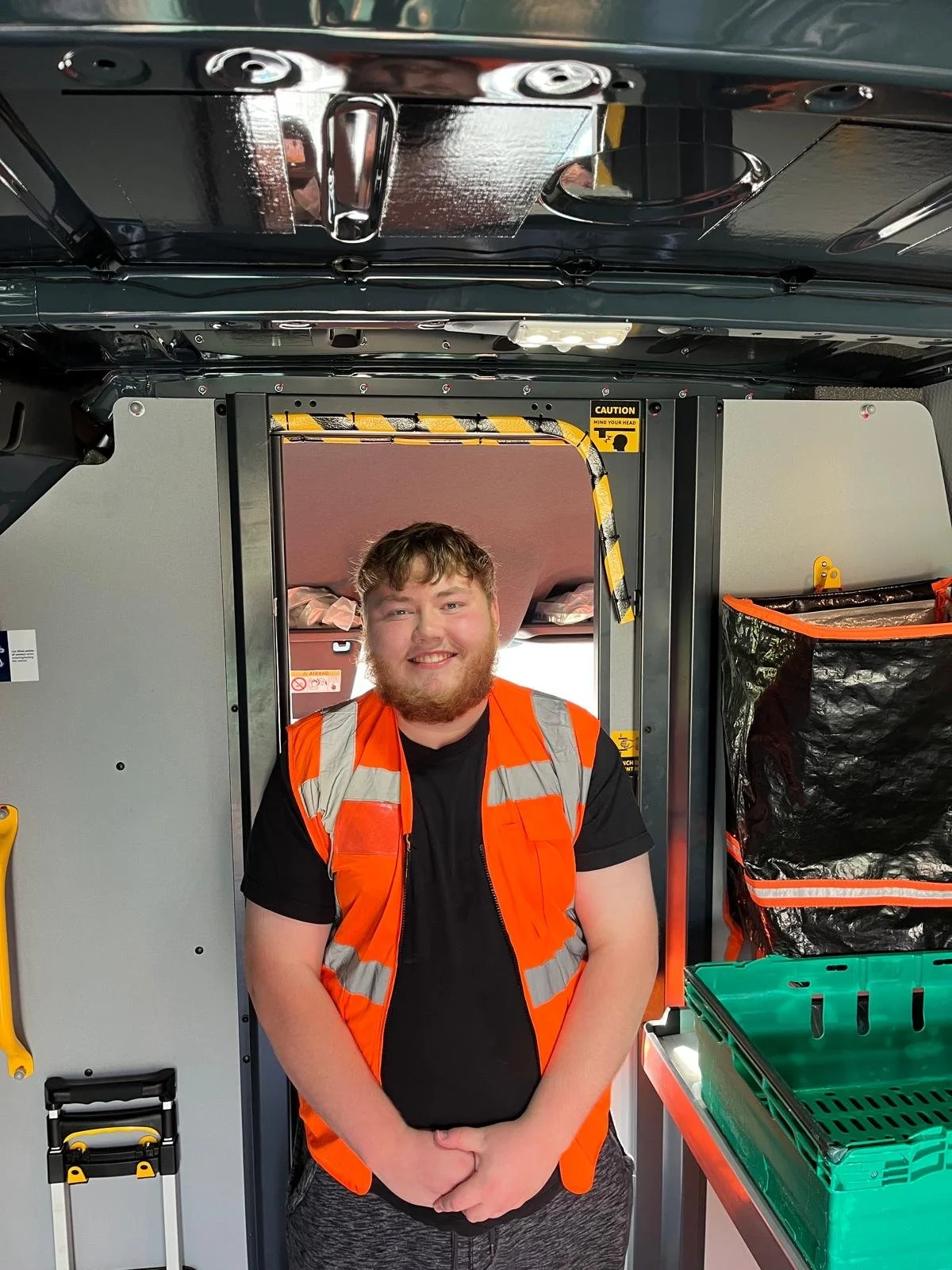 A smiling man wearing a black t-shirt and an orange reflective safety vest standing inside a vehicle, a delivery truck or van, with various storage compartments and equipment visible around him.