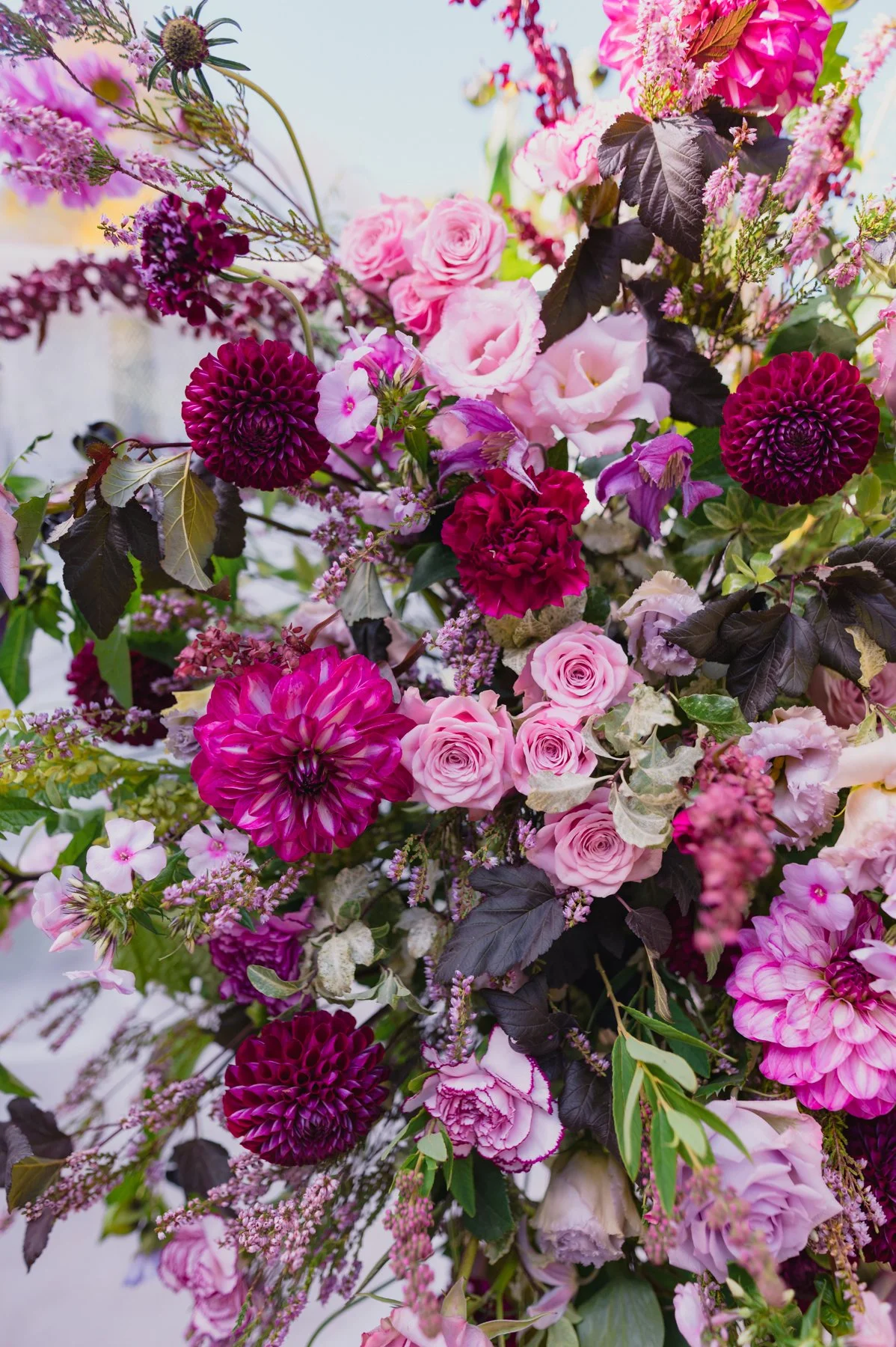 Un bouquet de fleurs colorées comprenant des roses roses, des dahlias violets, et d'autres fleurs roses, avec des feuilles vertes et foncées.