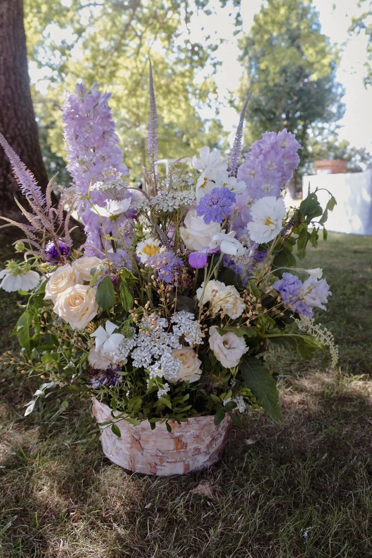 Un arrangement floral dans un pot en bois, composé de roses blanches, de lavande violet clair, de fleurs blanches et violettes, avec un fond de arbres et ciel en plein air.