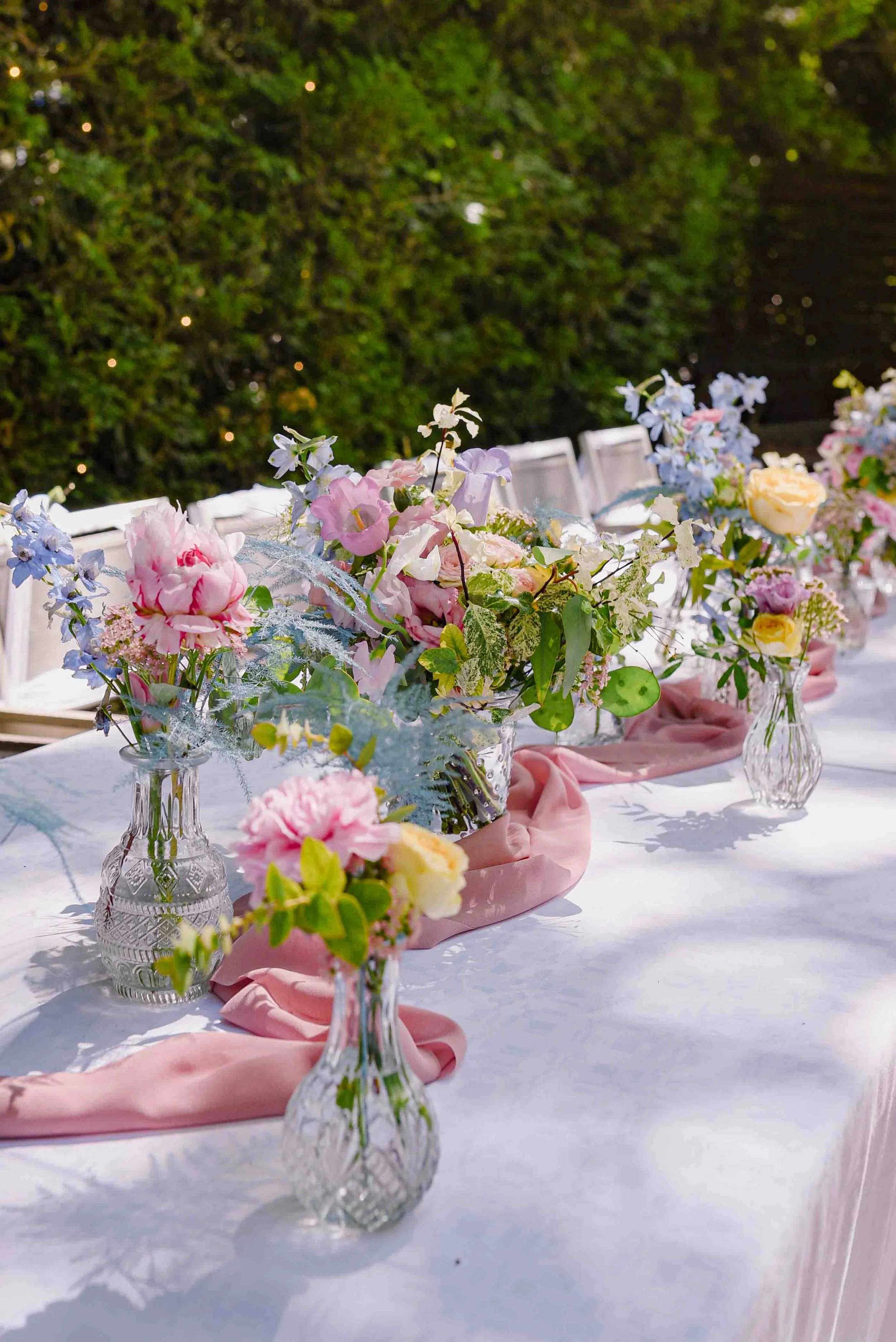 Table décorée avec des vases en verre contenant des arrangements floraux de différentes fleurs pastel, entourés d'un tissu rose pâle, dans un cadre extérieur avec un mur végétal en arrière-plan.