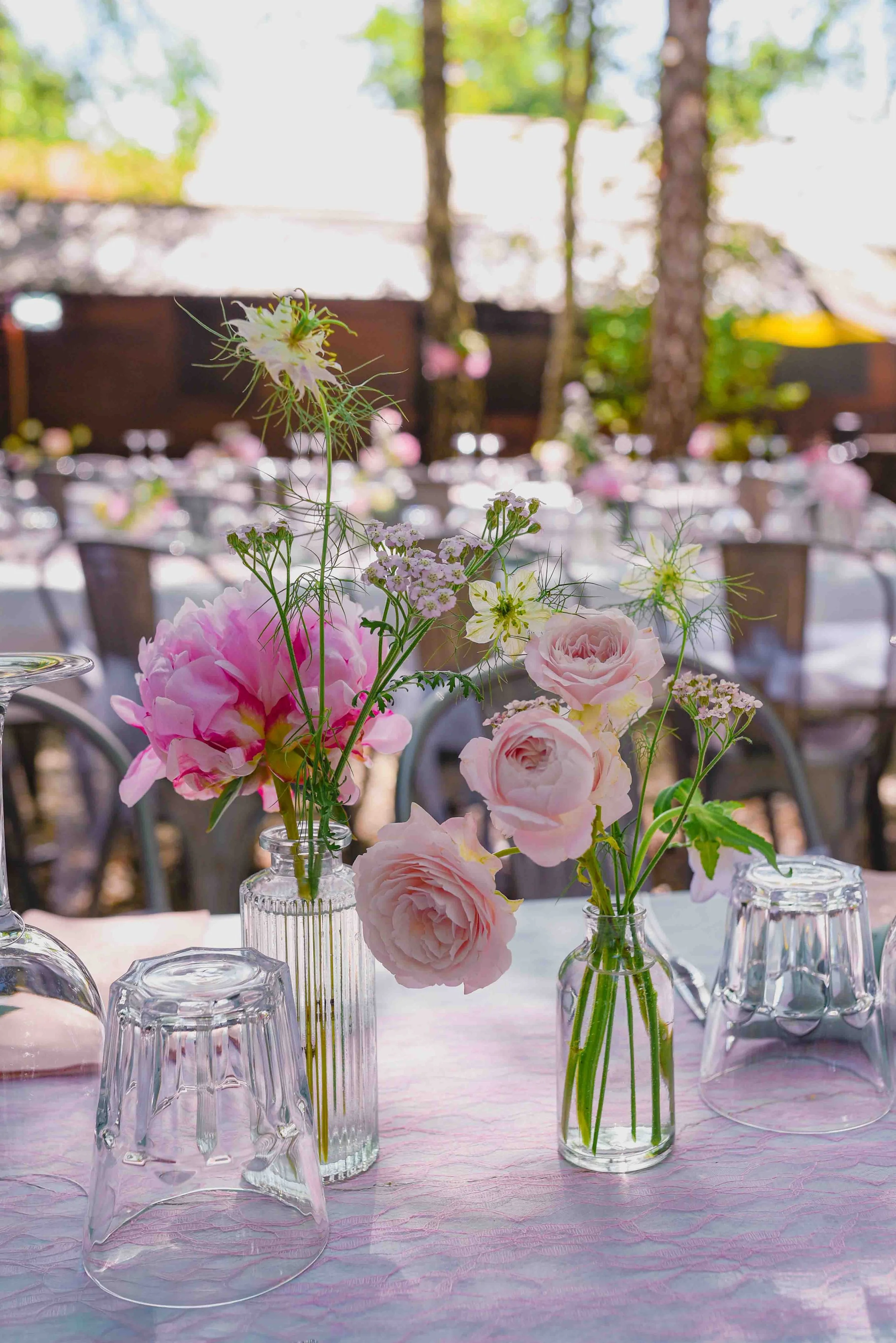 Vases avec des fleurs pastel, table décorée pour un événement en plein air, arbres en arrière-plan.