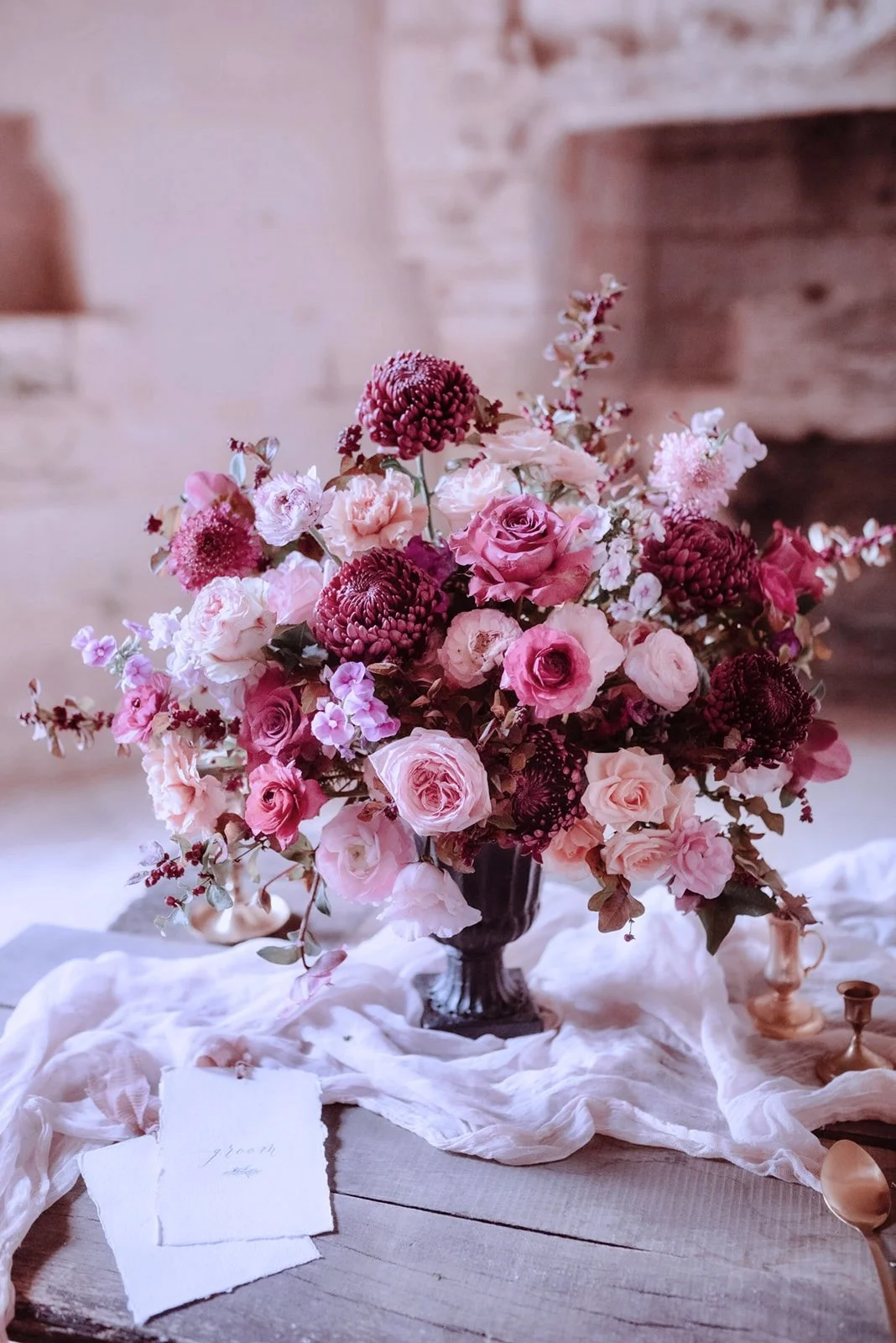 Un bouquet de fleurs roses et rouges dans un vase noir, posé sur une table en bois avec un tissu blanc doucement froissé, dans une ambiance chaleureuse et rustique.