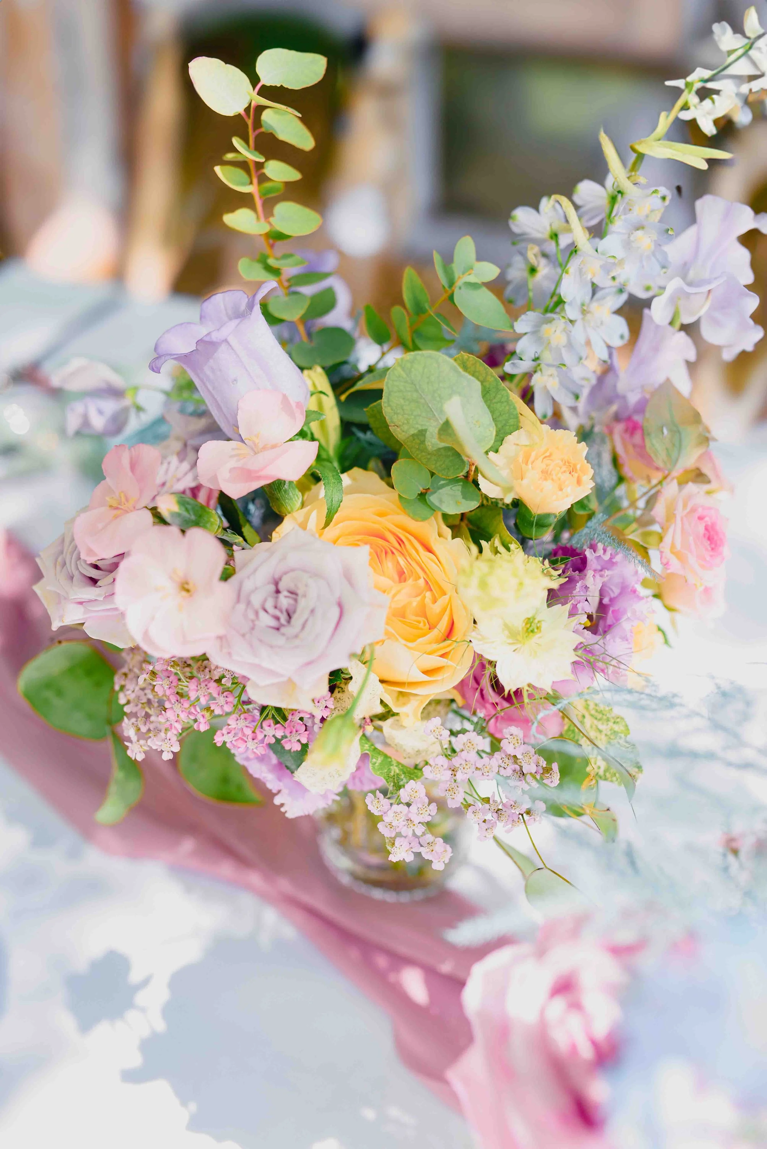 Un bouquet de fleurs pastel dans un vase rose, comprenant des roses, des pivoines, des fleurs de lavande, et des feuillages variés, placé sur une surface blanche avec une ombre douce.