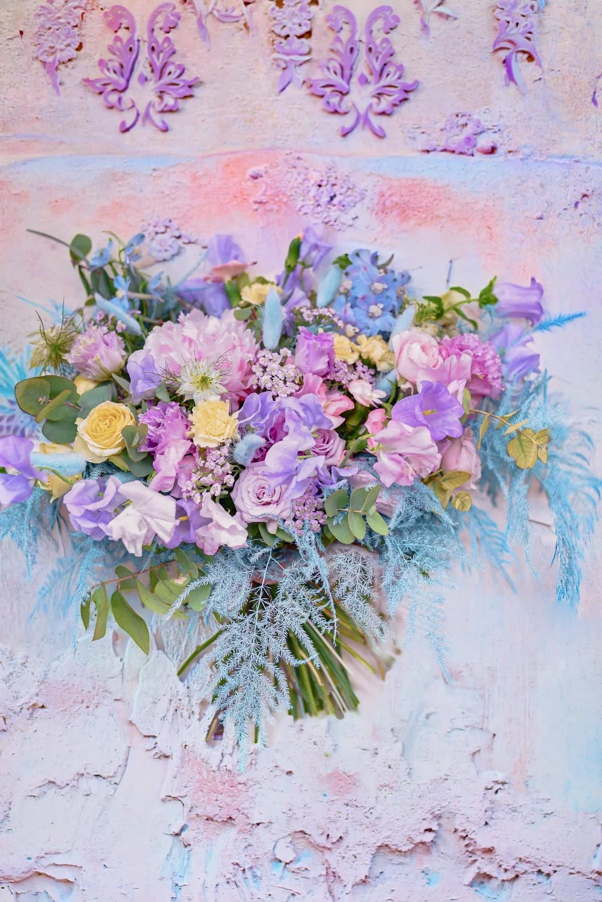 Un bouquet de fleurs colorées composé de roses, lis, arborvitae, et autres fleurs pastel, posé contre un mur peint en pastel avec des decorations en relief.