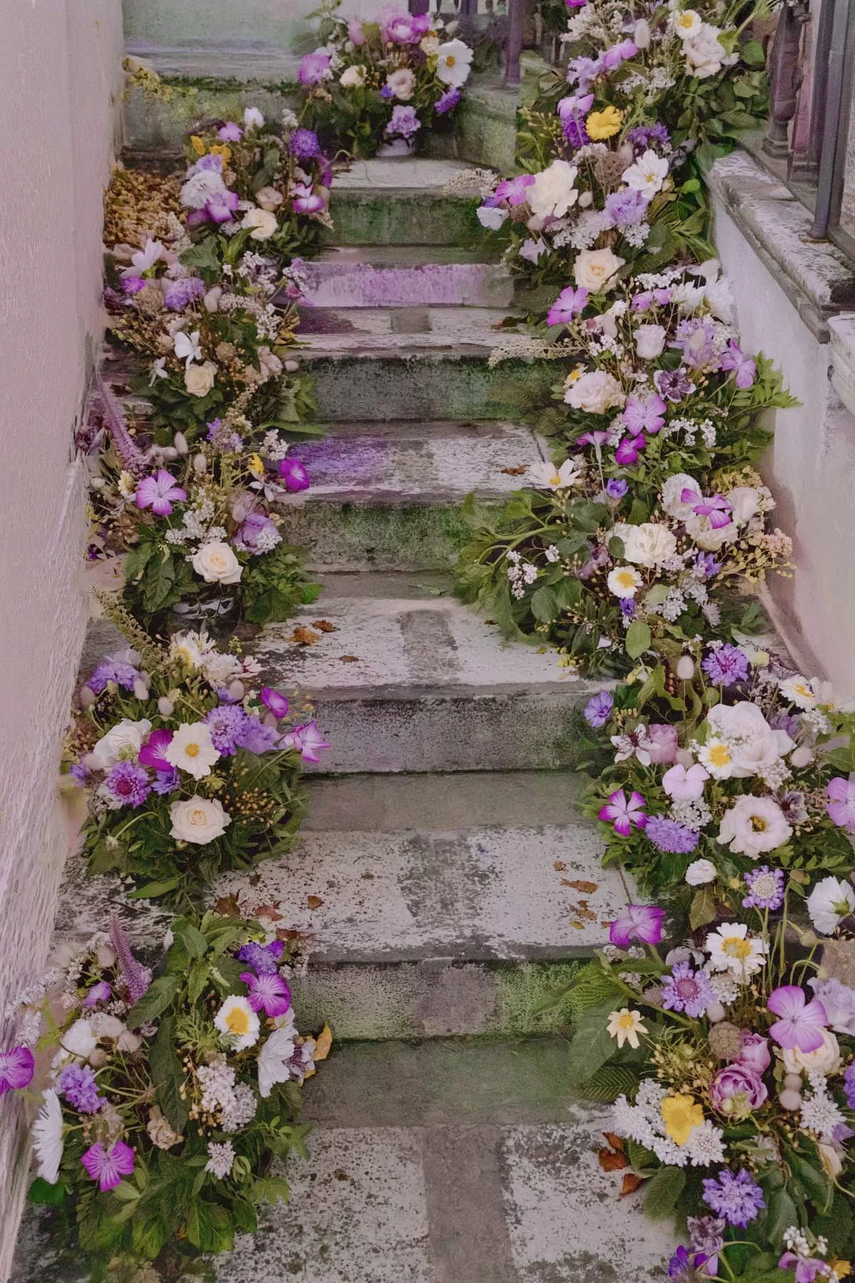 Escalier en pierre ancienne décoré de guirlandes de fleurs colorées, notamment roses, marguerites, et autres fleurs violettes et blanches.