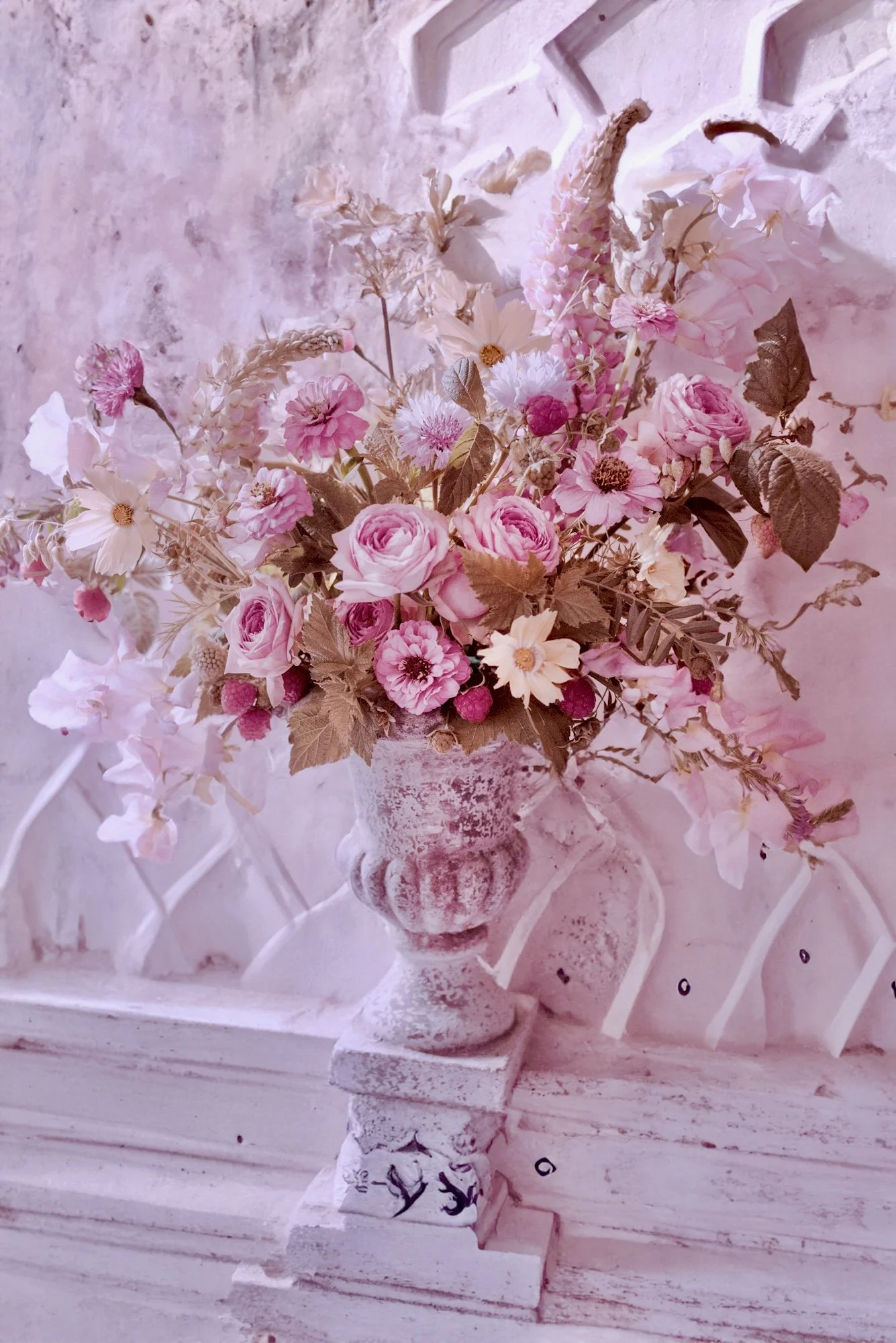 Un arrangement floral avec des roses et autres fleurs roses dans un vase blanc, posé sur une surface en blanc, avec un fond en pierre blanche et un motif en diagonale.