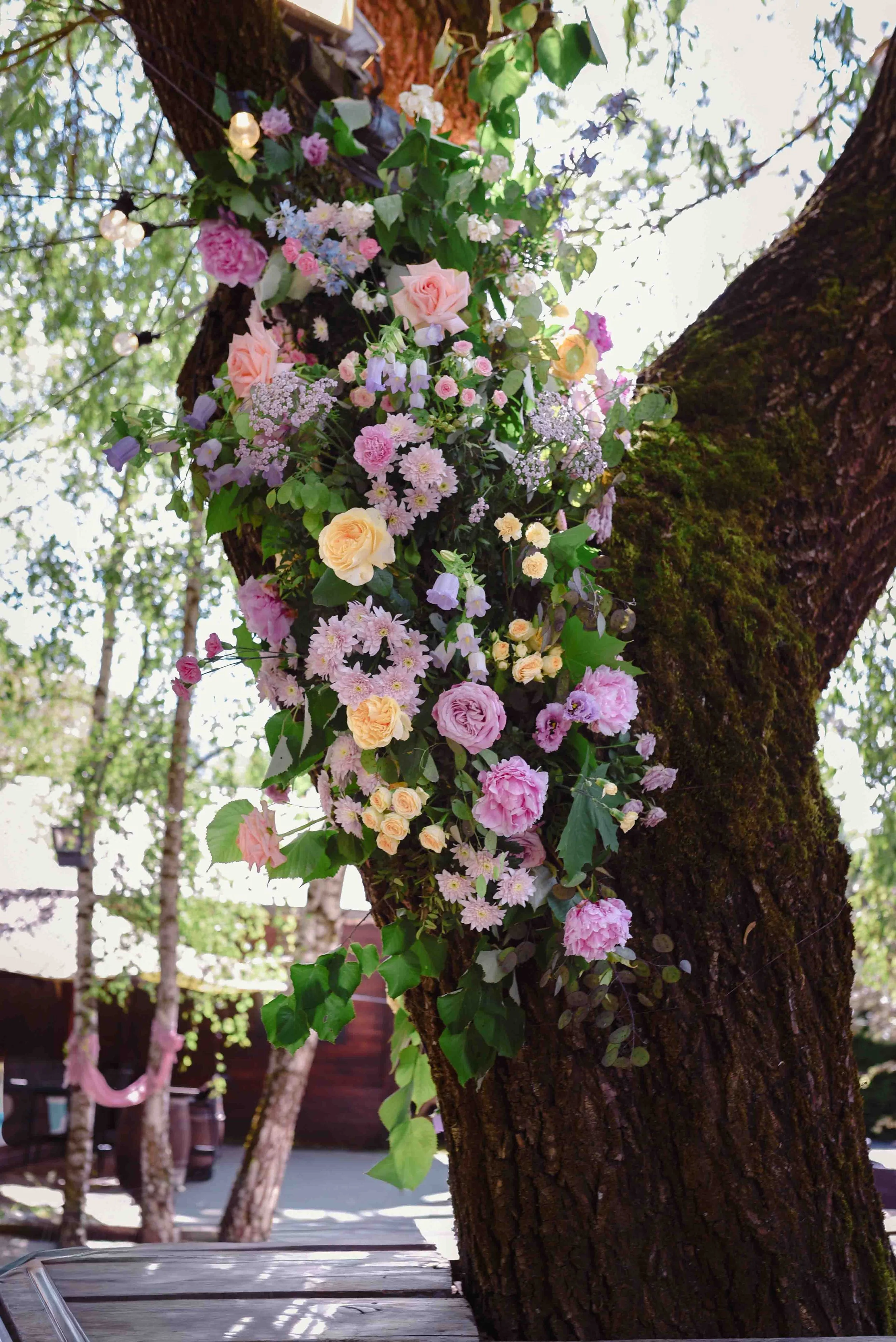 Arbre décoré avec des fleurs de différentes couleurs, notamment roses, violettes et blanches, avec des lumières à ampoules suspendues dans un espace extérieur.