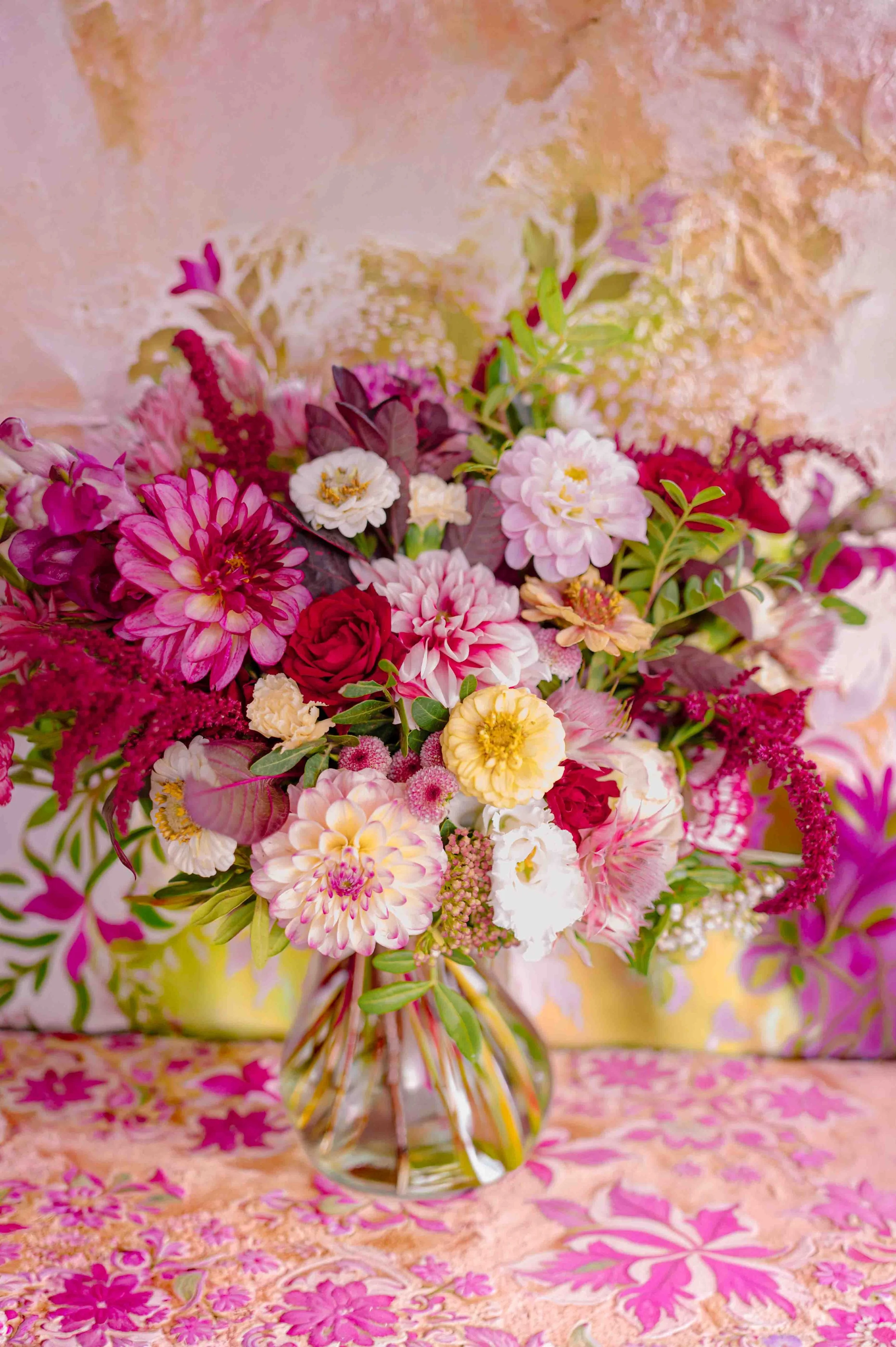 Un bouquet de fleurs colorées dans un vase en verre, placé sur une surface avec un tissu à motifs floraux roses et jaunes, avec un fond pastel pink et or