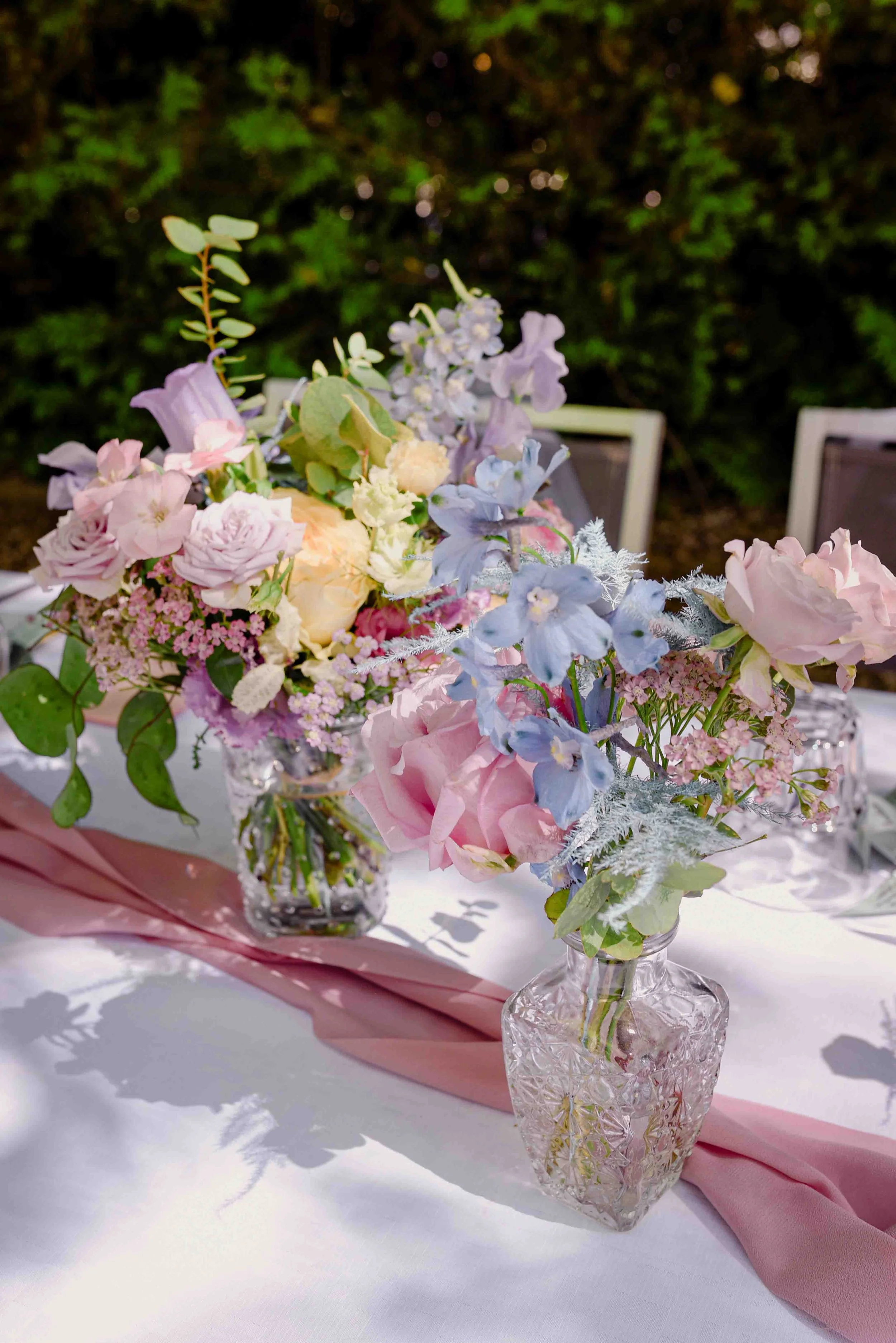 Un bouquet de fleurs dans un vase en cristal avec une nappe rose et une table blanche, en extérieur, fond d'arbres et de feuillages.