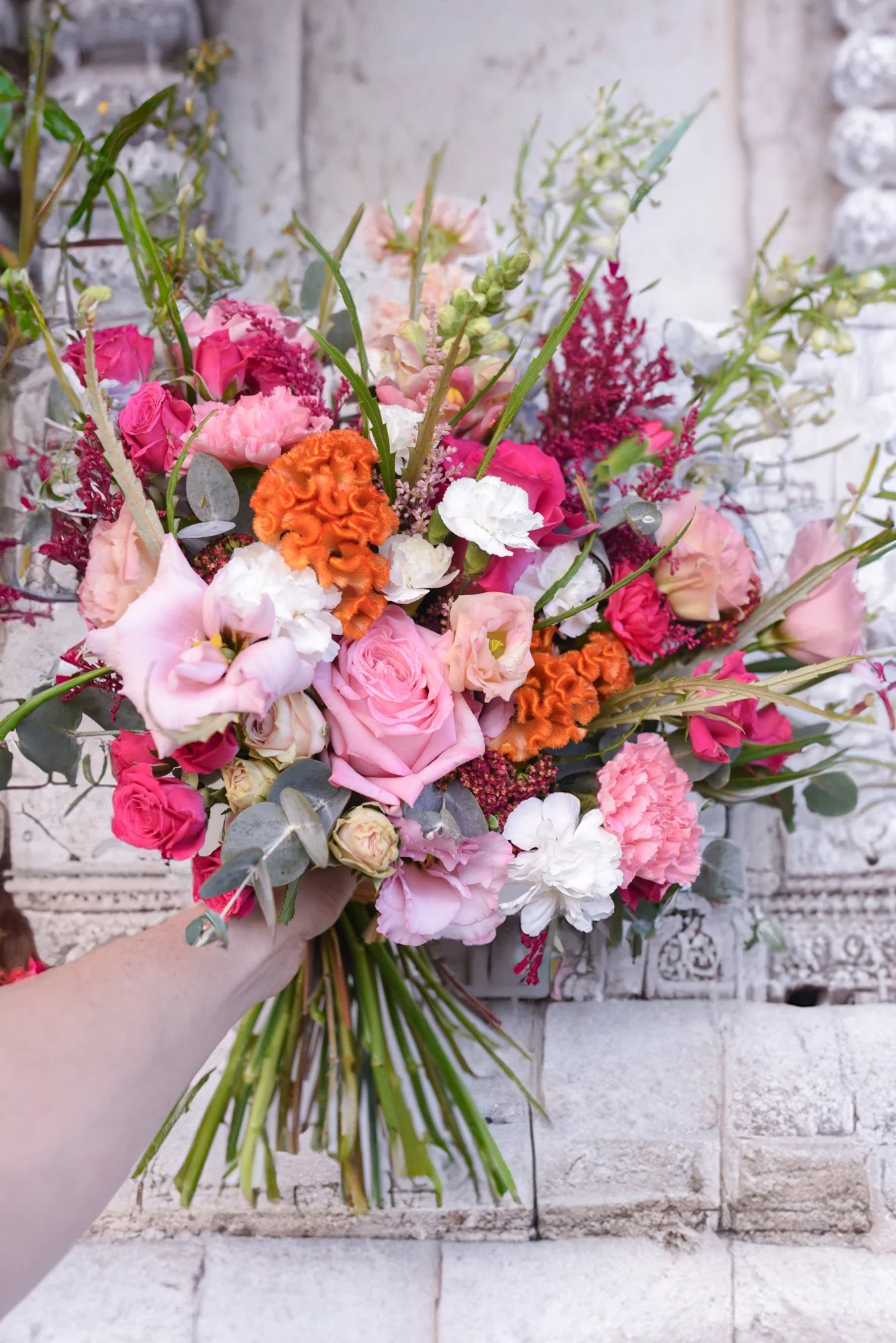 Un bouquet de fleurs colorées avec des roses roses, des fleurs orange, des lis blancs et d'autres fleurs dans des tons roses et blancs, tenu par une main, devant un mur en pierre.