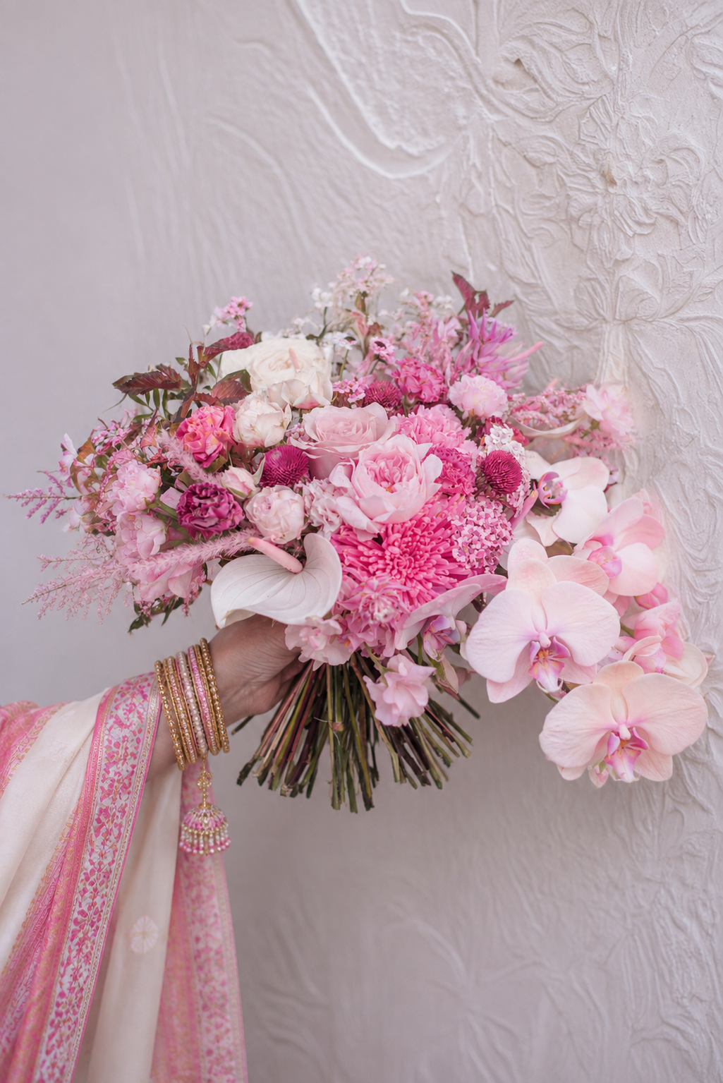 Un bouquet de fleurs roses, comprenant des roses, des orchidées et d'autres fleurs, tenu par une mariée portant des bangles en or et sari indien tissu rose et beige avec des détails dorés, en face d'un mur blanc texturé.
