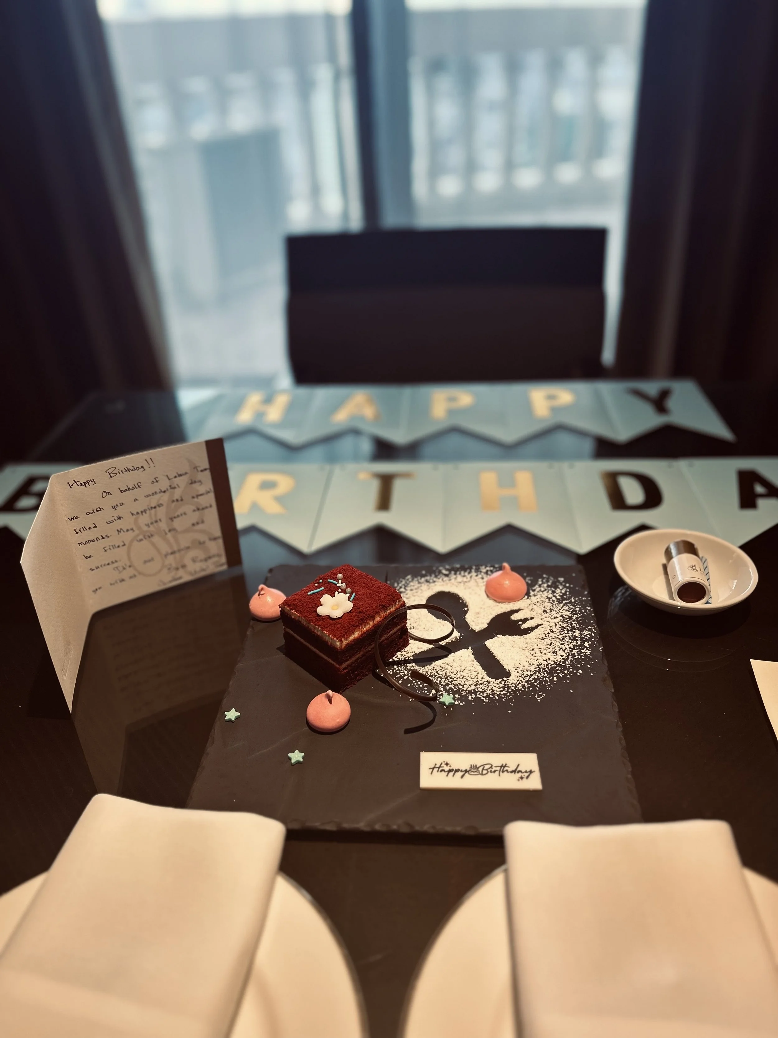 Person lying on a bed with white sheets, looking out a large window at a scenic view of the sea and distant cityscape during daylight.