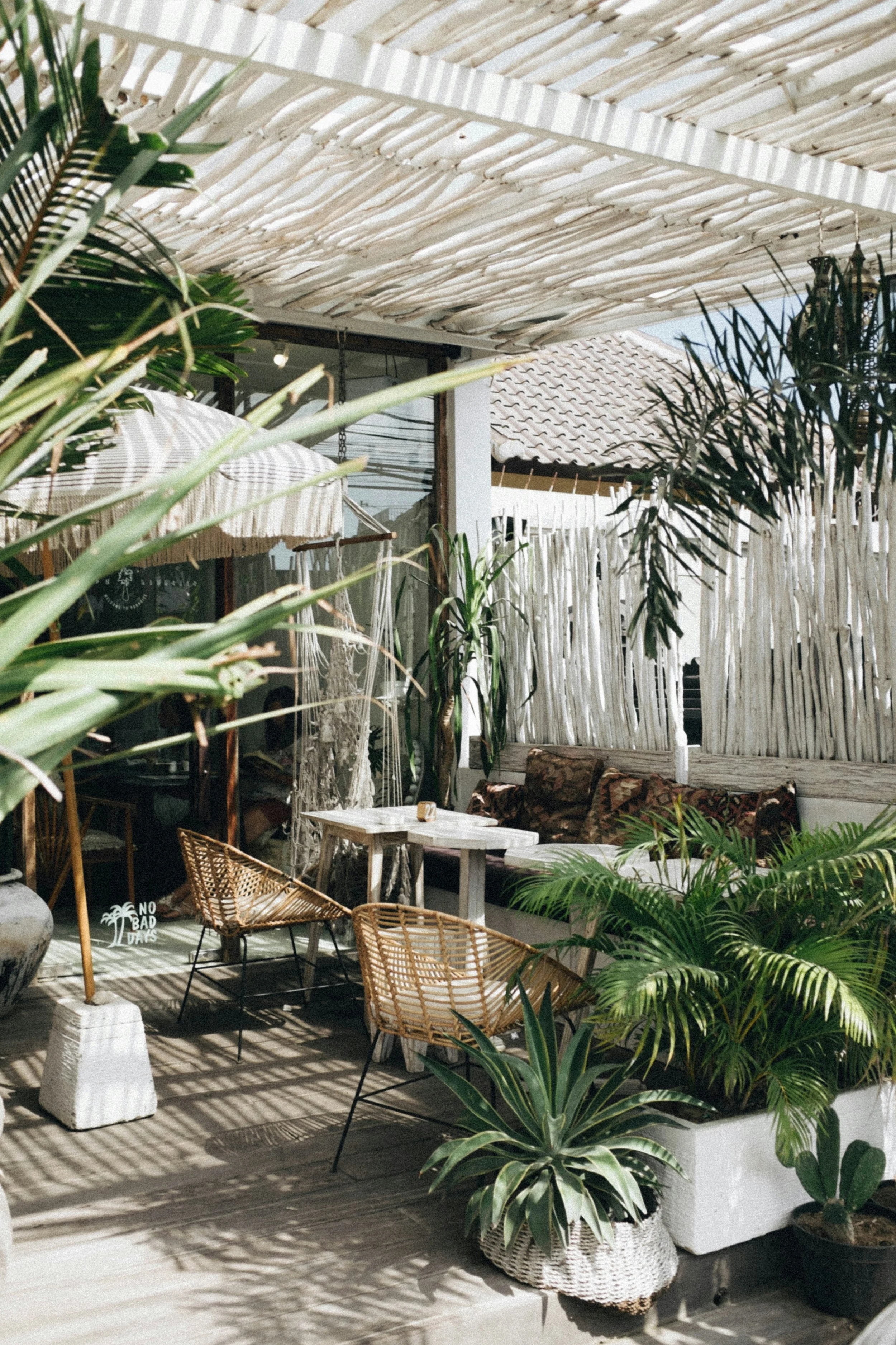 Outdoor patio with wicker chairs, white wooden table, large potted plants, bamboo roof, and decorative pillows on a bench.