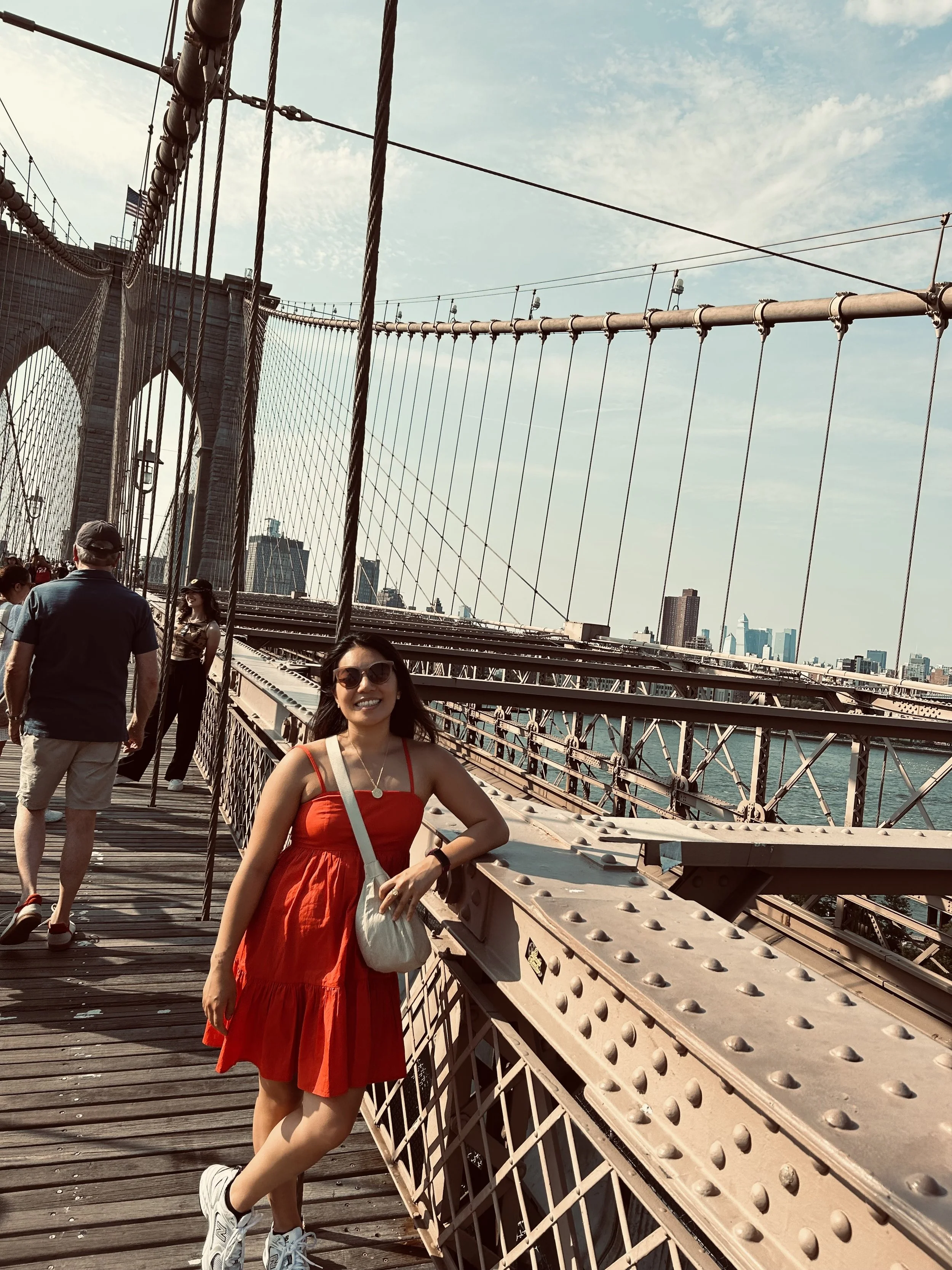 Person in a hat standing in an infinity pool with a view of the ocean, surrounded by lush greenery and a pier in the background.