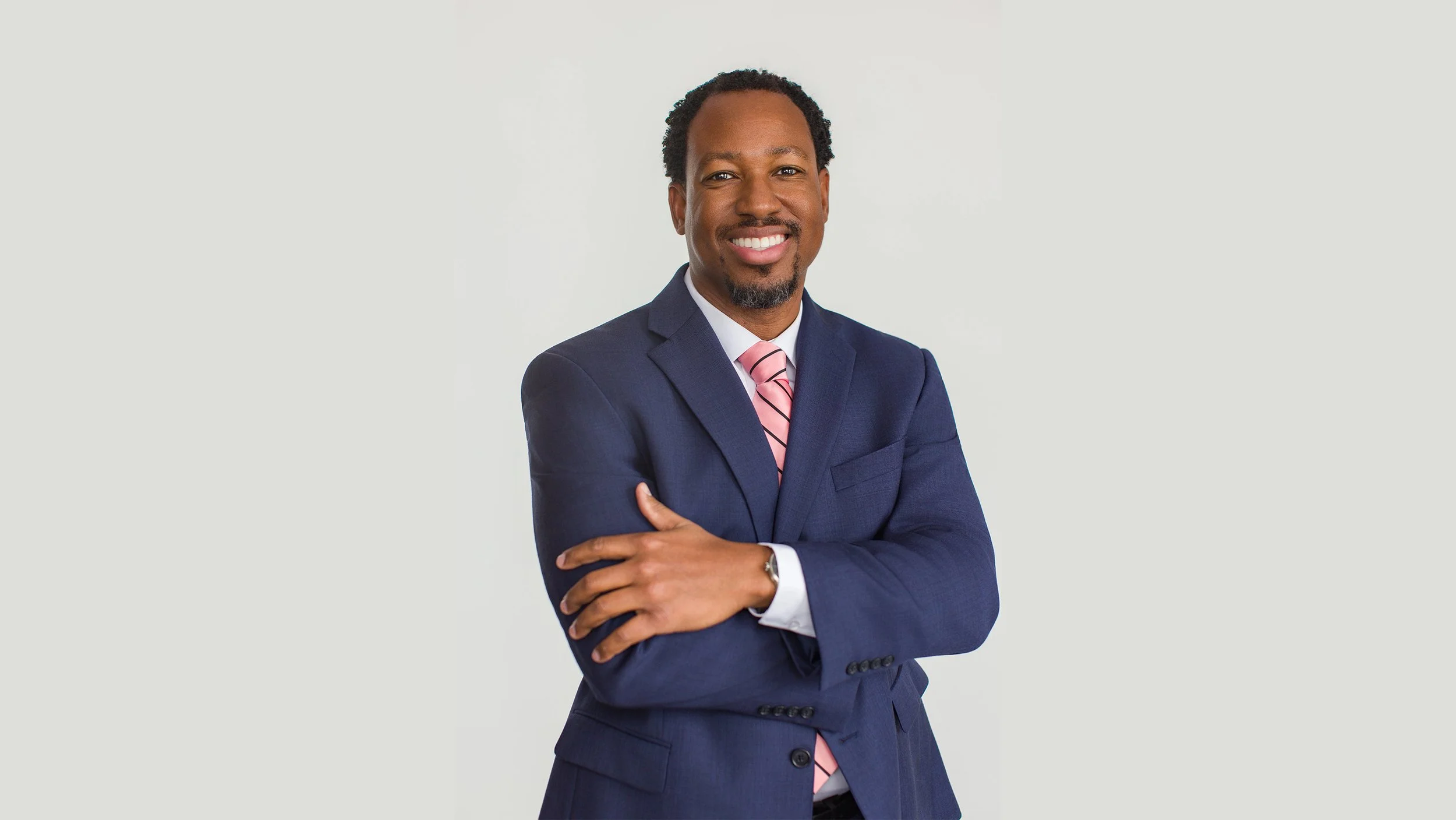 Confident businessman in a navy suit and pink tie smiling with arms crossed against a clean white studio background.